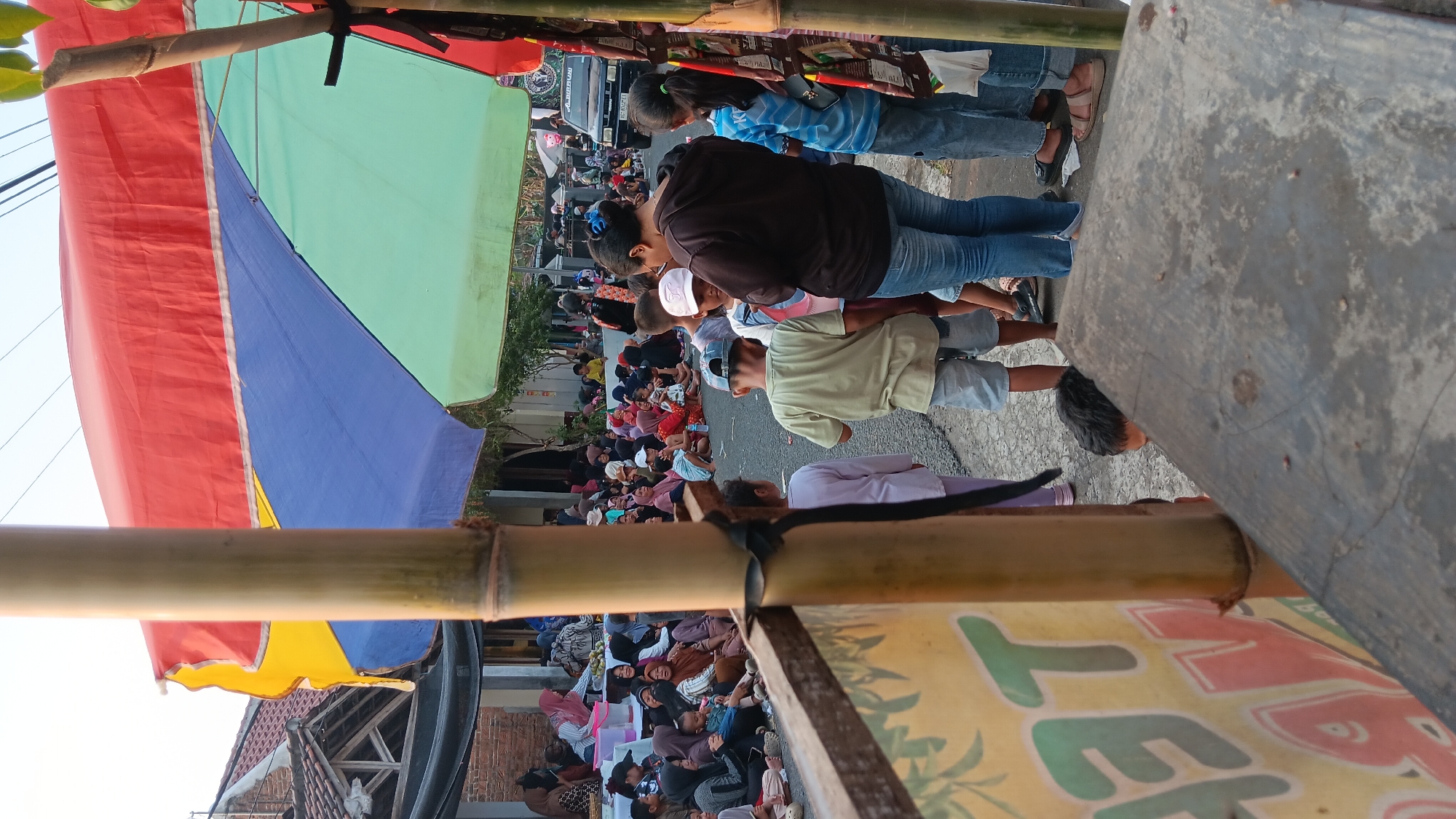 This is a vertical, eye-level, medium shot of a street scene in Cerme, Indonesia, captured from behind a bamboo pole with a banner on its left. A large, multi-colored umbrella, red, blue, and green, shades a gathering of people. In the foreground, the bamboo pole and a concrete surface are prominent. A group of children and adults are standing and sitting on the street. On the right side of the frame, a black vehicle is parked on the side of the road. The backdrop consists of buildings with red tile roofs and some trees. The overall impression is of a community gathering or event.
