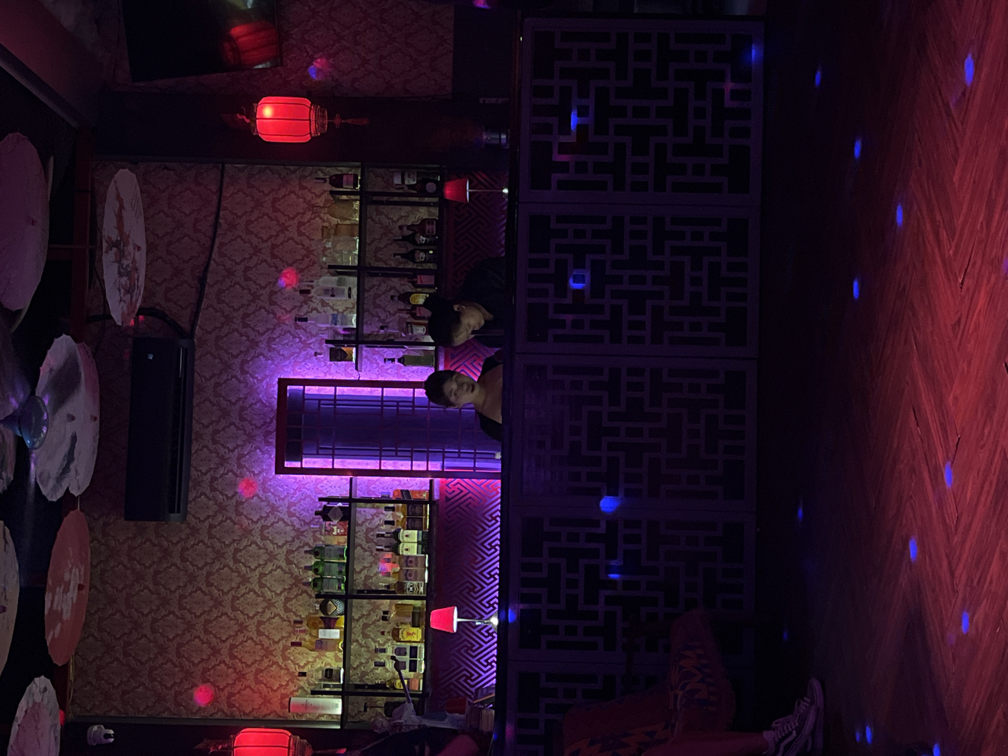 The image depicts the interior of a bar or restaurant in Đà Nẵng, Vietnam. The space features a bar counter with an intricate geometric design on its facade. Shelves behind the bar are stocked with liquor bottles. A red lantern hangs from the ceiling, and red and purple lighting sets the mood. Two individuals are visible behind the bar. The flooring is wooden with a pattern of planks and illuminated by blue lights. Other decorative elements include paper umbrellas adorning the wall, and patterned wallpaper.