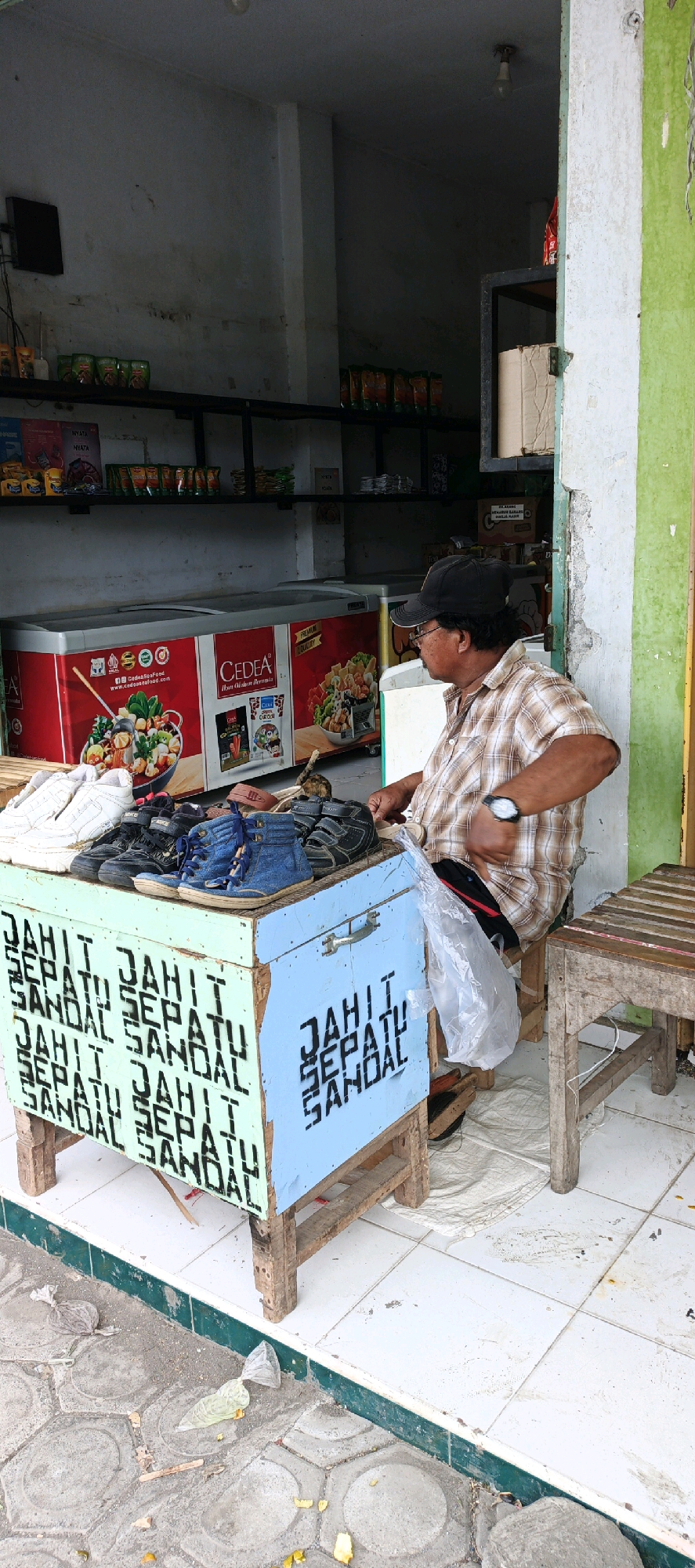 In Kasiyan, Indonesia, a middle-aged man wearing a checkered shirt, black baseball cap, and glasses sits on a stool next to a stall. The stall, constructed from light blue and green painted wood, displays various shoes and sandals. The side of the stall is stenciled with the phrase "JAHIT SEPATU SANDAL," which translates to "shoe and sandal repair." In front of the stall are two large red refrigerators with images of food items. The man is holding a plastic bag and appears to be working with a shoe. The ground around the stall is paved with hexagonal concrete tiles and scattered debris. Shelves behind the stall are stocked with packaged goods. The overall scene depicts a small business operating outdoors.