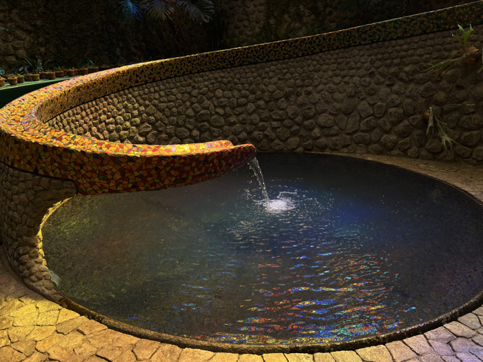 The image displays an architectural water feature located within an enclosed space in Naucalpan de Juárez, Mexico. A dark, circular water basin occupies the lower half of the frame, with water actively flowing into it from an elevated spout. The surface of the water exhibits colorful, fragmented reflections and concentric ripples originating from the point of impact. The water spout is the terminus of a prominent, elevated, curved structure, resembling a serpent or wave, which is entirely covered in a vibrant mosaic of irregularly shaped tiles primarily in shades of yellow, orange, red, and brown. This mosaic-covered structure originates from the upper left and curves downwards towards the water basin. A textured wall constructed from irregular grey and brown stones forms the background, curving behind the water feature. Small potted plants are arranged on a ledge along the upper left section of this stone wall, and additional green foliage is visible on the wall's upper right portion. The ground immediately surrounding the water basin is paved with irregularly shaped, yellowish-tan stones. The scene is illuminated by artificial light sources, creating areas of highlight on the mosaic and surrounding paving, while casting the background stone wall in shadow. No individuals are present.