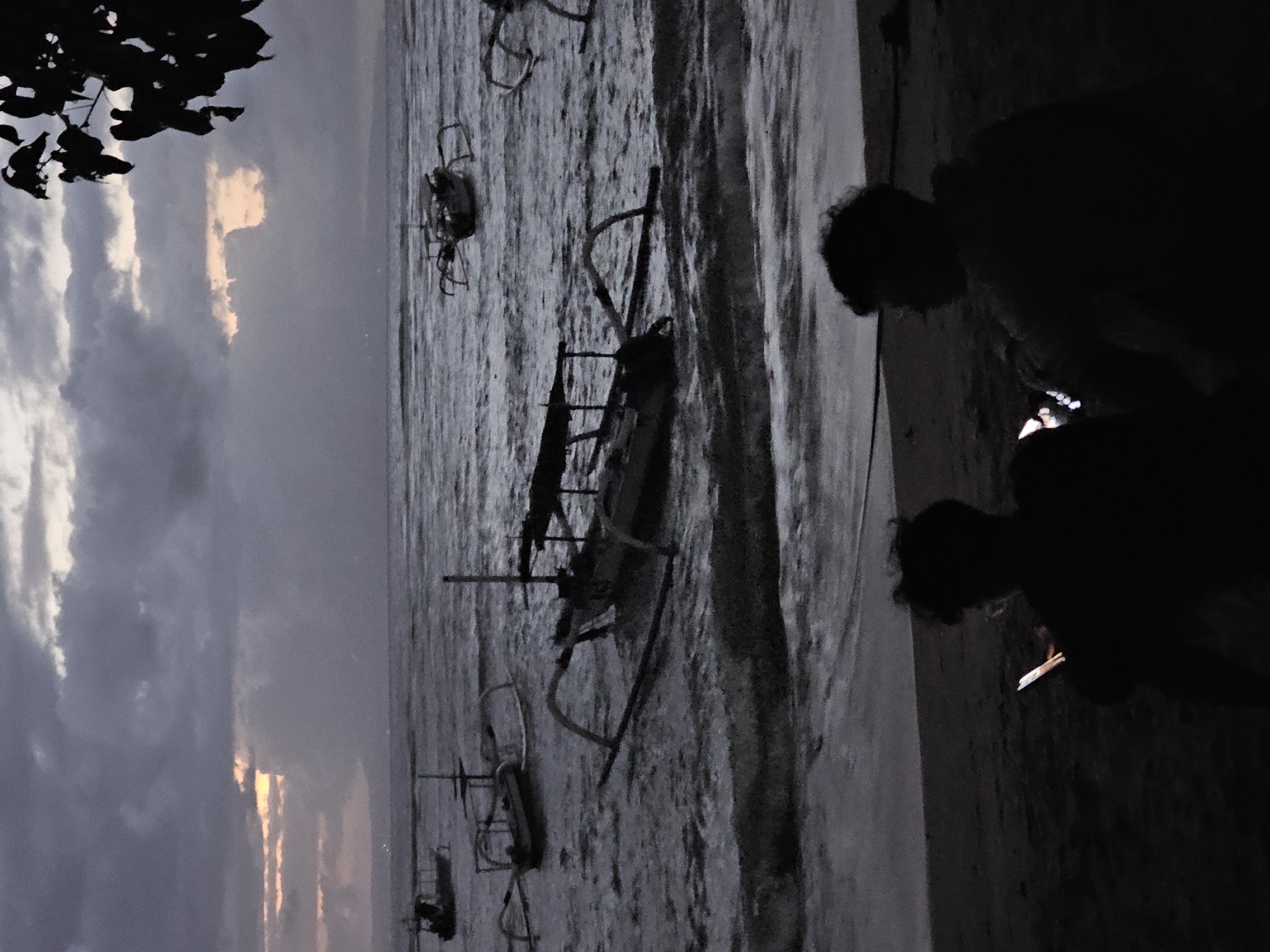 The image shows a beach scene in Denpasar, Indonesia, at twilight or dusk. Several outrigger boats are pulled up onto the sand. Two silhouetted figures stand on the beach; one appears to be looking at a phone. The sky is cloudy with a hint of sunset.