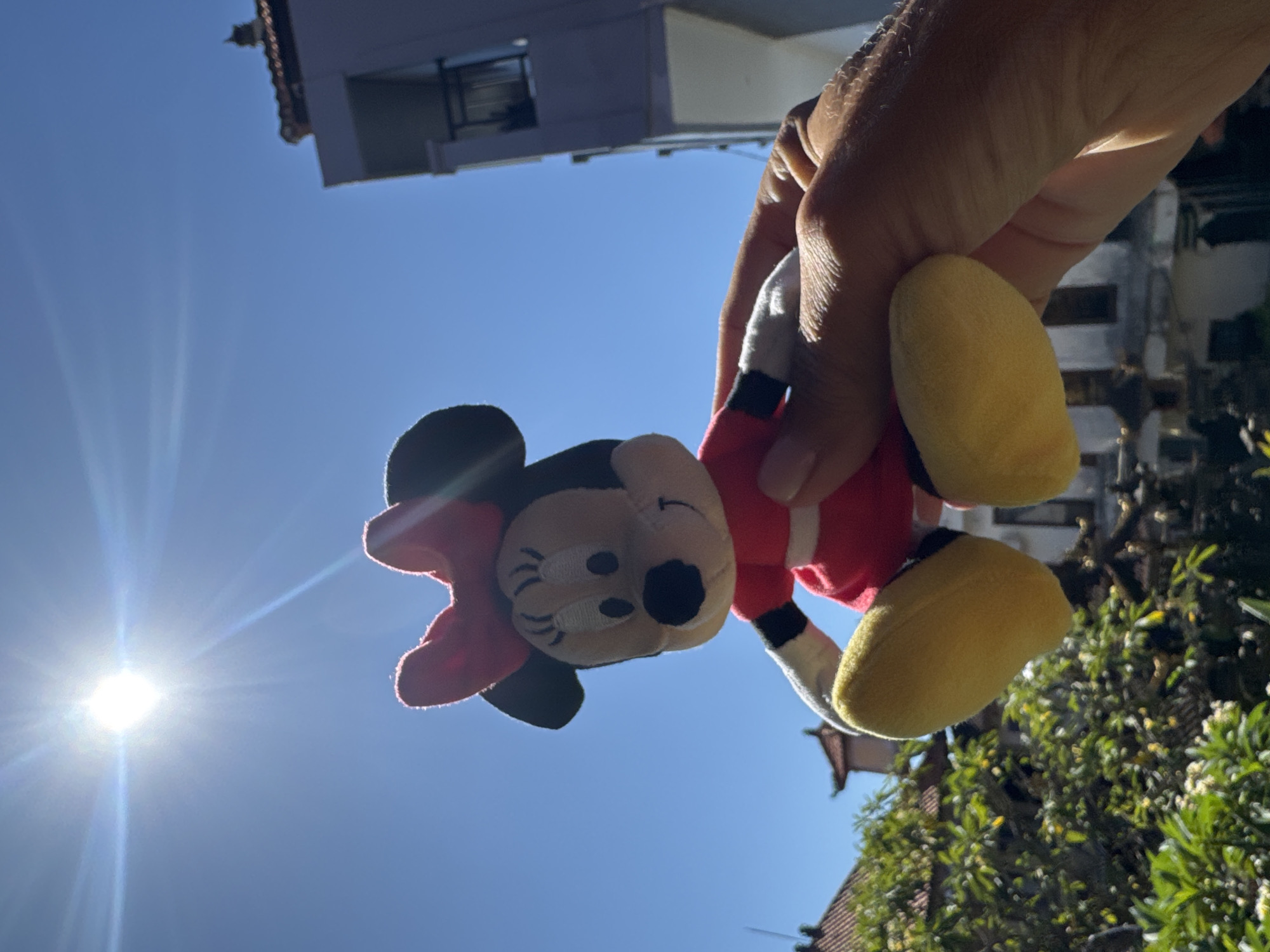 A hand holds a Minnie Mouse plush toy against a bright blue sky with a sun flare. The background features architectural elements, including a building with a tiled roof, a modern building with a balcony, and green trees, indicative of a tropical environment. The image was likely taken in Kuta, Indonesia.