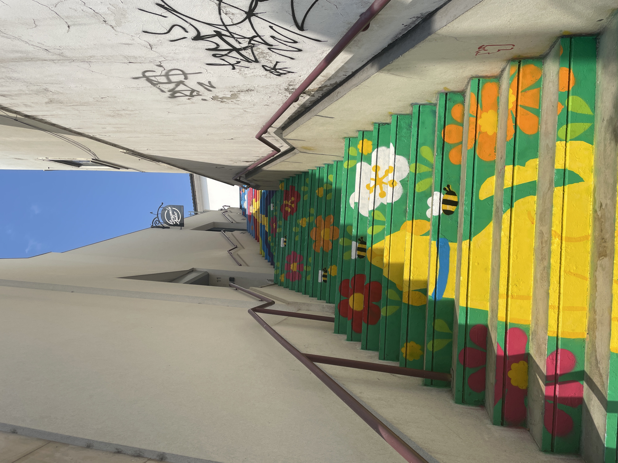 The image depicts a vibrant, art-covered staircase situated in the city of Águeda, Portugal. The stairs are painted a bright green and adorned with colorful floral motifs and bumblebee illustrations, creating a cheerful and lively aesthetic. The staircase is flanked by two buildings, one on the left with a smooth, light-colored facade and a building on the right with a rough, textured, and graffitied wall. A metal railing runs along both sides of the stairs, providing support. The sky above is a clear, bright blue. A sign for "Segundo Andar" is visible on the left building, indicating a second floor. The overall scene is a public space transformed by street art, likely a pedestrian passageway or a decorative architectural feature.
