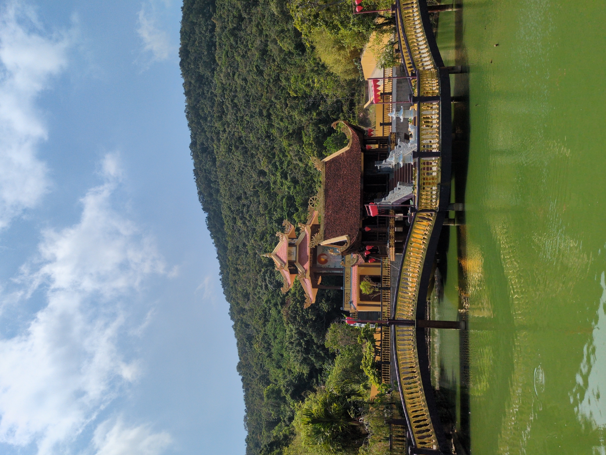 A scenic view of a traditional Vietnamese temple complex is seen, built next to a lush, green mountainside and beside a body of water. A bridge, decorated with intricate gold railing, connects the temple buildings to the land. The complex features several structures with red-tiled roofs, detailed architectural designs, and red lanterns. The sky is light blue with scattered white clouds. The green of the foliage on the mountain contrasts with the green of the water. The location is likely in or near the city of Phú Quốc, Vietnam, as evidenced by the traditional temple design and the tropical scenery.