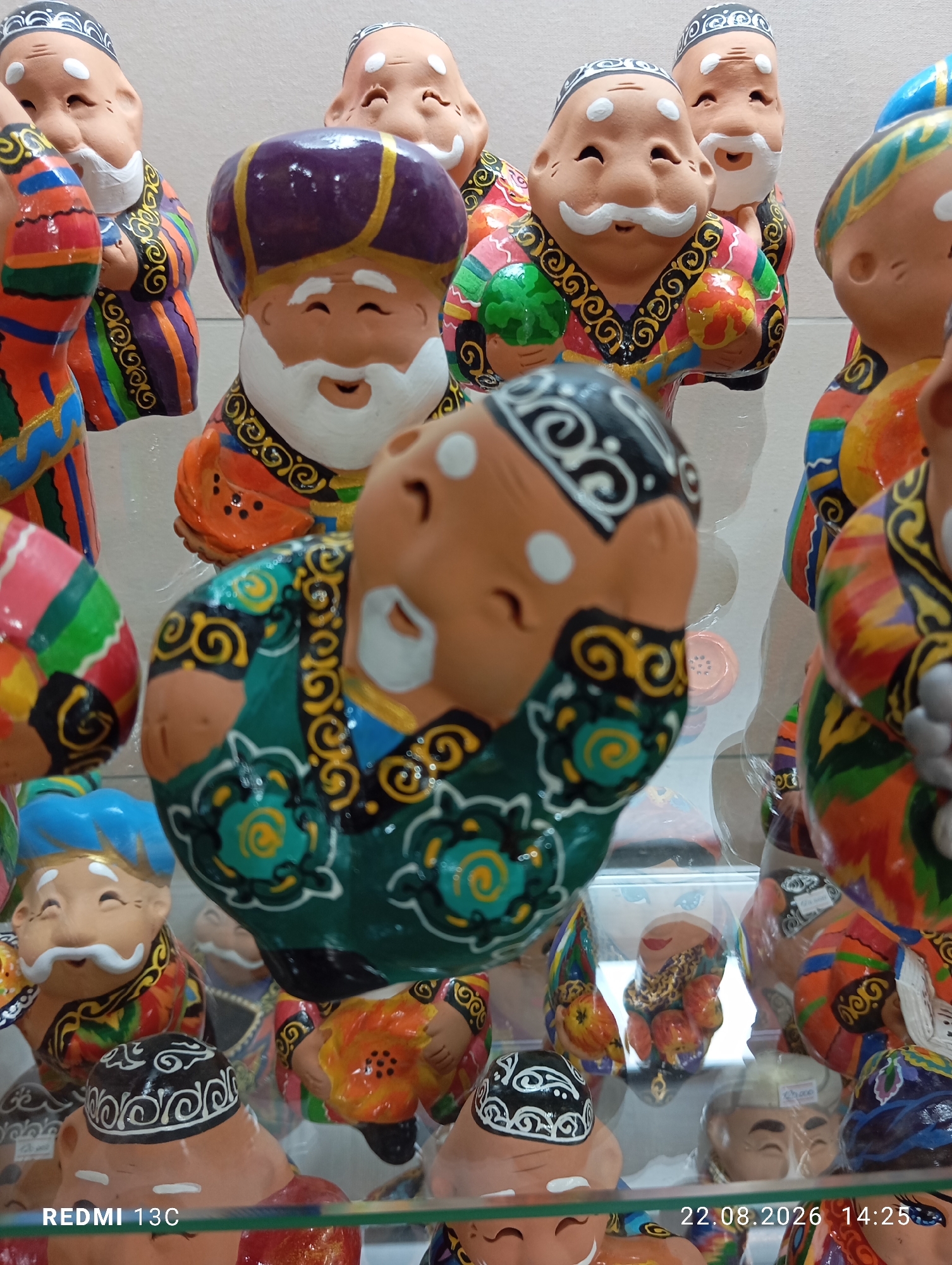 In a retail setting in Amirsay, Uzbekistan, a collection of vibrantly colored ceramic figurines are displayed on glass shelves. The figurines depict stylized male figures, likely representing traditional Uzbek men, adorned in ornate robes with intricate patterns in shades of orange, blue, green, red, and yellow. Many wear traditional skullcaps or turbans. Their faces are simplified, with white beards and mustaches, and eyes often depicted as closed or semicircular, giving them a cheerful or serene expression. Some figurines are holding small objects, such as green and red spheres. The central figurine is tilted forward, appearing to be in motion or a dynamic pose. The background is a plain, light color, and a white price tag is visible on one of the figurines in the lower right corner. The lower portion of the image shows the "REMI 13C" text on the left and a date stamp "22.08.2026 14:25" on the right, indicating a timestamp of the photograph.