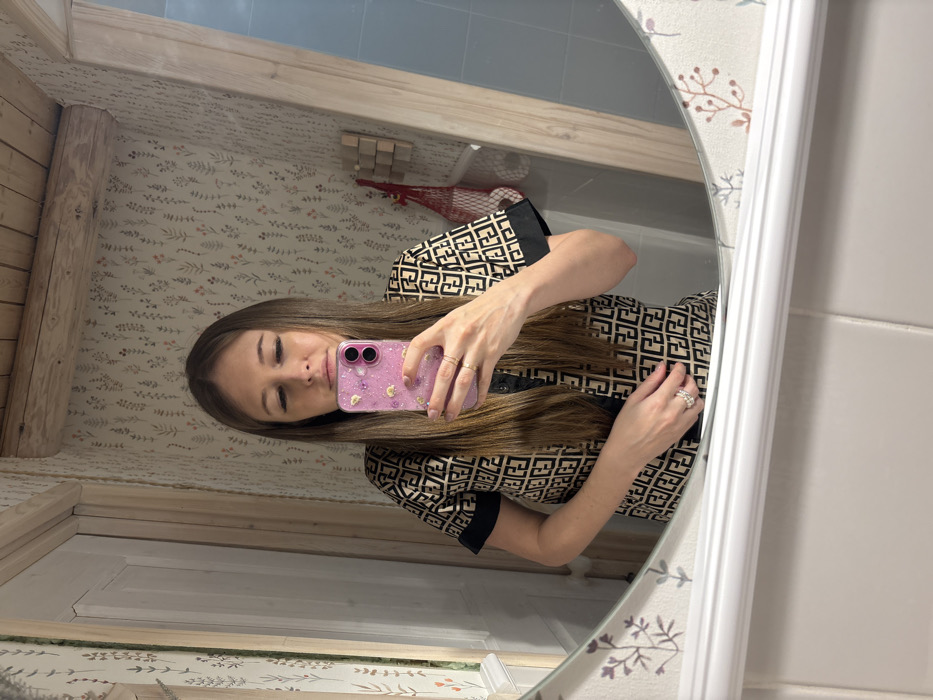 A woman is captured in a mirror selfie, reflecting her head and upper torso. She has long, straight brown hair and wears a dress or top featuring a repeating black geometric pattern, resembling a stylized "F" or "FF" monogram, on a light tan/beige background, with black trim on the sleeves and placket. She holds a smartphone in a translucent pink case adorned with glitter and small golden charms, positioned to capture her reflection. Rings are visible on both her hands; simple bands on her left and a multi-band diamond or crystal ring on her right. Her expression is calm, with her eyes appearing downcast or slightly closed.
The mirror has a white, fluted frame on its right side. The background reflected in the mirror reveals an indoor setting, likely a bathroom or dressing area. Features include light-colored wallpaper with a subtle botanical/floral pattern in muted green and reddish-brown tones, and exposed light-colored wooden beams or trim, suggesting a rustic aesthetic. In the upper right of the reflection, a small wall-mounted wooden object with multiple square components is visible, alongside a partially seen red mesh bag. To the right of the mirror's frame, a white tiled wall is apparent, and deeper in the reflection, blue/grey tiles and a potential bathtub or sink area can be glimpsed. The location context provided is Lezhnevo, Russia.