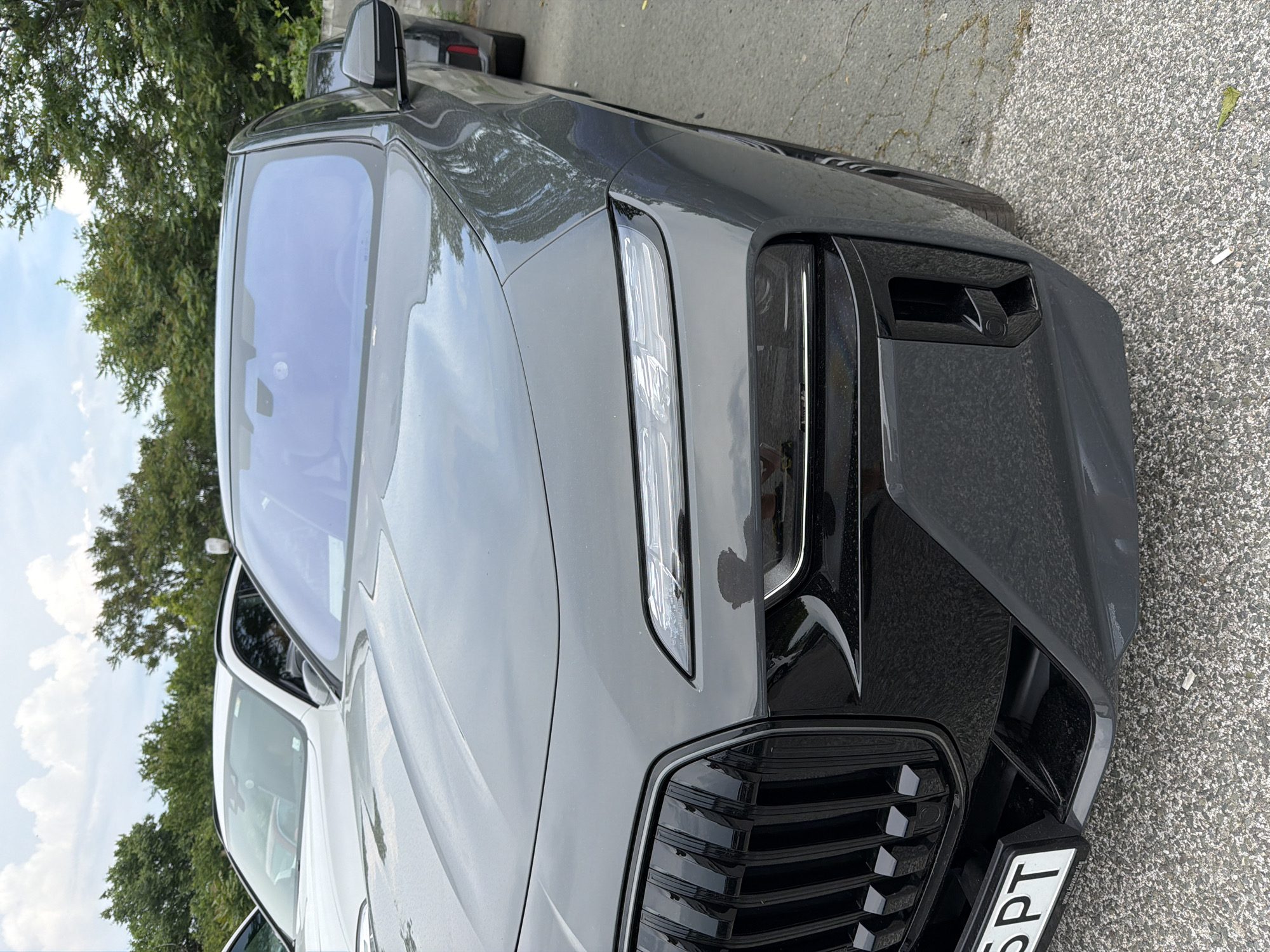 A dark grey (charcoal) modern SUV or crossover, likely a BMW, is parked outdoors in Burgas, Bulgaria, under bright, partly cloudy skies. The image is taken from a low angle, focusing on the front and front-right side of the vehicle.
The car features a distinctive front-end design with slim, horizontally-oriented LED daytime running lights positioned high on the fender line. Below these, the main headlights appear integrated into the bumper area, contributing to a sleek and aggressive look. A large, prominent, and somewhat squared-off kidney grille with vertical slats dominates the front, characteristic of the brand. The bumper also includes air intakes and a small, square-shaped sensor or camera housing on the lower right side.
Reflections of green trees and the blue sky with white clouds are visible on the car's glossy dark grey paintwork and the expansive front windshield. A black, angular side mirror is also visible. In the background, lush green trees and a clear blue sky with scattered clouds suggest a daytime setting with pleasant weather. The car is parked on textured, grey asphalt pavement. Another lighter-colored vehicle is partially visible behind the main subject. A portion of the front license plate is visible, showing the letters 'SPT'.