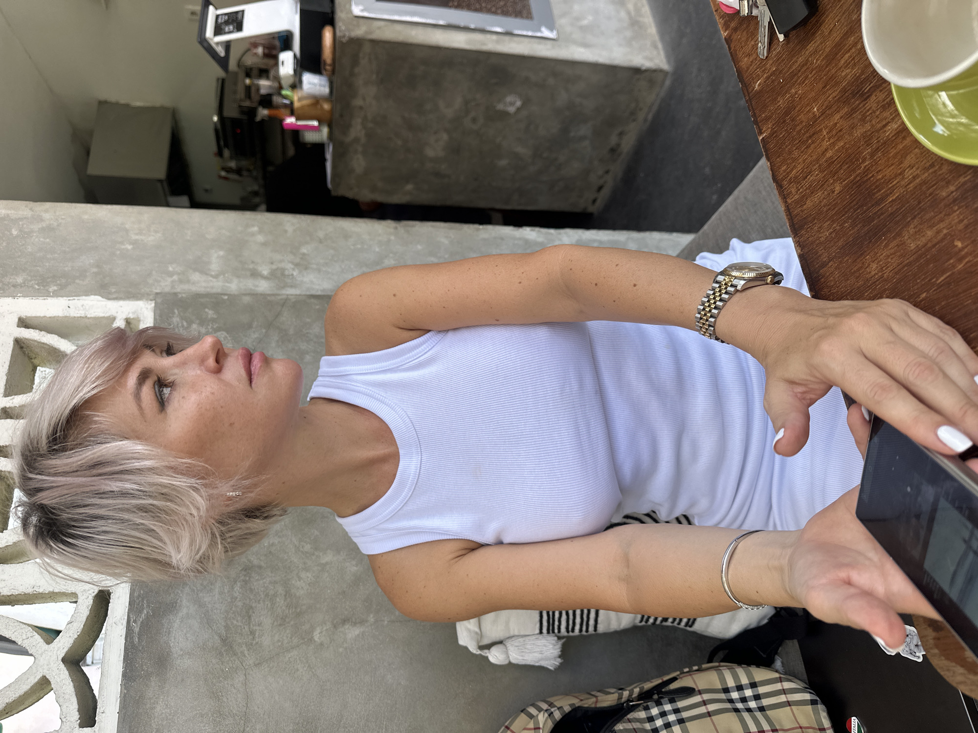 A medium shot, eye-level photograph captures a Caucasian woman with short, blonde hair with pink undertones, looking to her right. She is wearing a white ribbed tank top and sits at a wooden table. On her left wrist, she wears a silver bracelet, and on her right wrist, a two-tone watch with a gold and silver band. Her hands are resting on the table, with her left hand holding a smartphone and her right hand hovering over it. White-tipped fingernails are visible on both hands. To the left of the woman, a plaid bag with a brown and tan pattern rests against a textured concrete wall. In the background, a concrete counter with various items, including a cash register, bottles, and a wooden cup holder with utensils, is visible. The lighting suggests an outdoor or semi-outdoor setting, possibly a cafe or restaurant, with a portion of a building visible to the right.