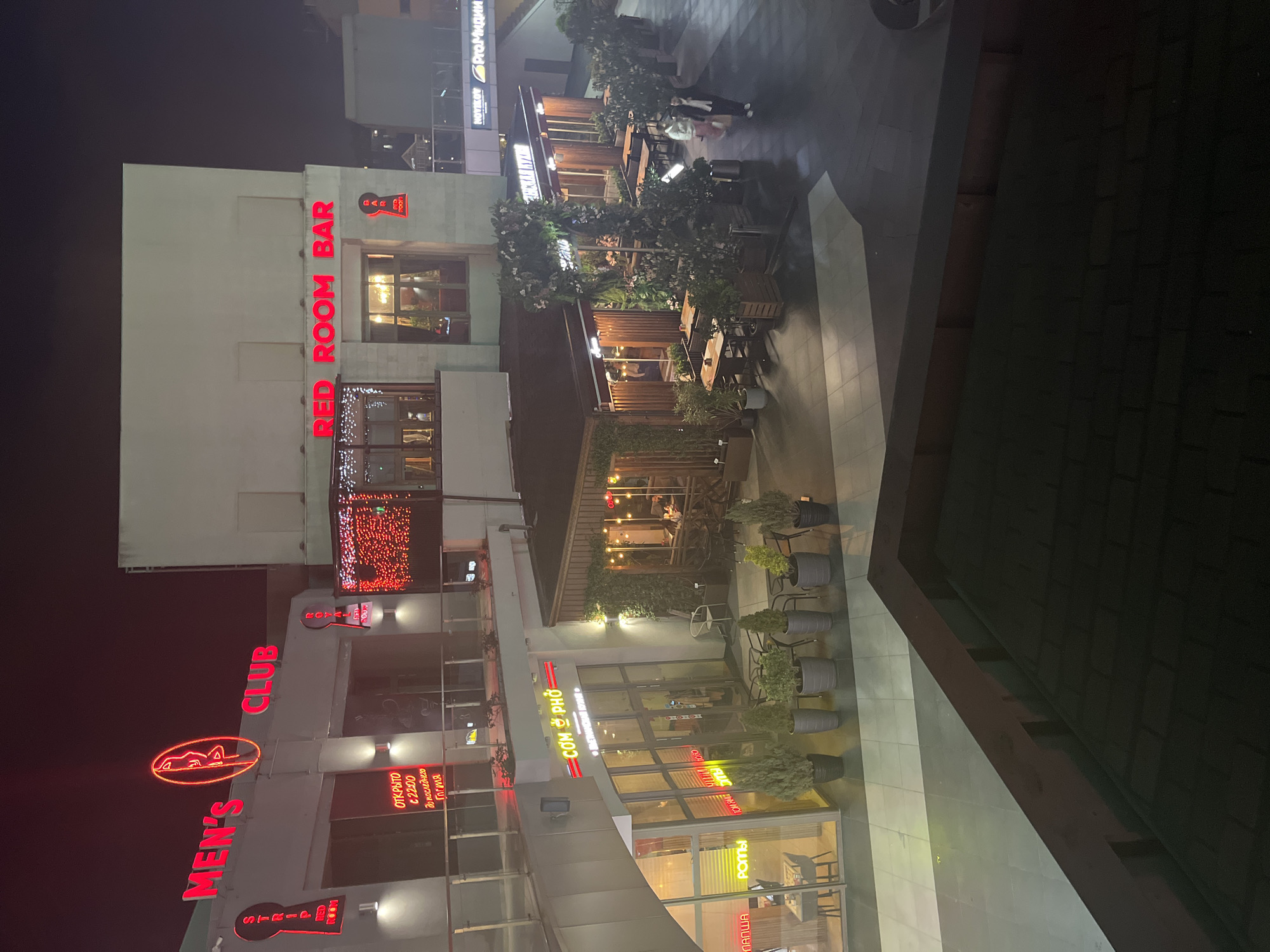 This is a nighttime shot of buildings in Sochi, Russia. The buildings are lit with neon signs and outdoor lighting. At the bottom left, there's a building with a neon sign that reads "MEN'S CLUB" and a neon silhouette of a reclining woman. Above that is "CLUB". Under it is "STRIP RED ROOM". On the same building, there is Russian text "ОТКРЫТО C 22:00 до последнего Гостя" which translates to "OPEN from 10 PM until the last guest". Next to it, "PAPSHA". On the building next to it is "COM O PHO" in red. The signs "TOM HA" "POME" and "ROTA" are visible. There is a building higher up with a neon sign that reads "RED ROOM BAR" in vertical red letters, with a small cartoon icon. Outdoor seating is seen in front of the buildings. Further up is "NOVIKOV ProМидии". There is an alley with tables and chairs visible, and two people are walking. The sky is dark, and the overall scene appears to be a lively nightlife area.