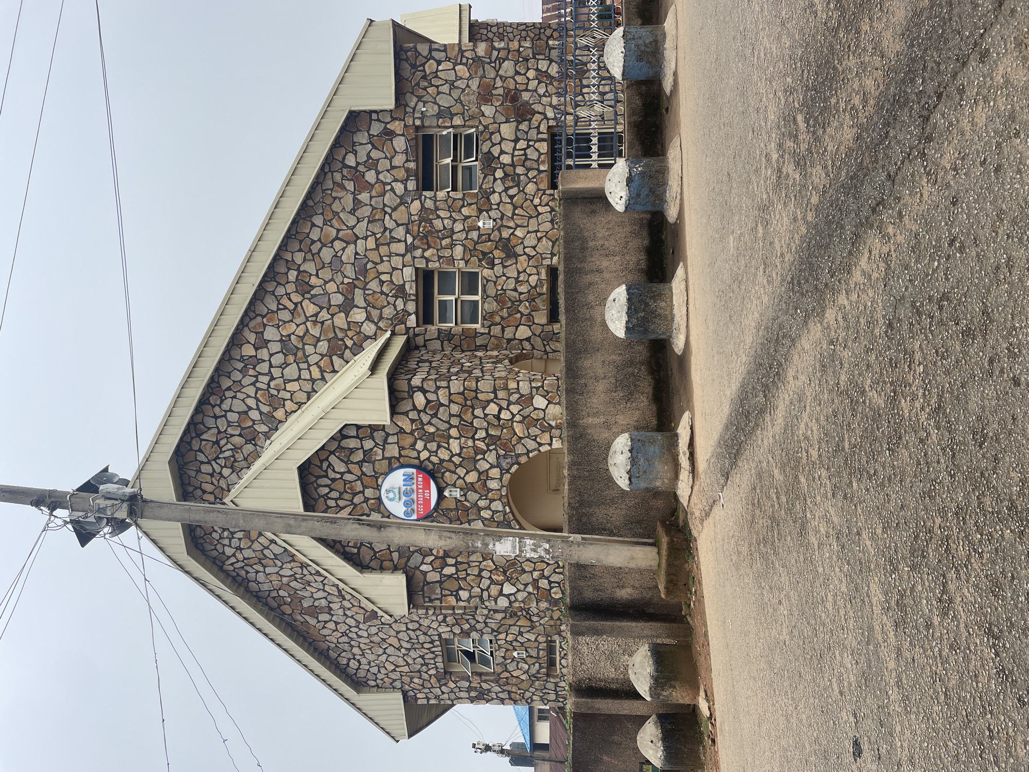 The image shows a building with a stone facade, likely a place of worship, in Jos, Nigeria. The building has a pitched roof with beige metal siding and a large arched entrance. A circular blue and white sign with the text "COCIN LCC BADIN KOWA JOS" is prominently displayed on the front of the building. A concrete utility pole with electrical wires and speakers stands in front of the building. Several concrete bollards are lined up along the sidewalk in front of the stone wall. The street in front of the building is paved with asphalt, and there is a shadow cast across it. The sky is overcast and the overall lighting is subdued. The scene appears to be an exterior view of a religious institution in a moderately urban or suburban environment.