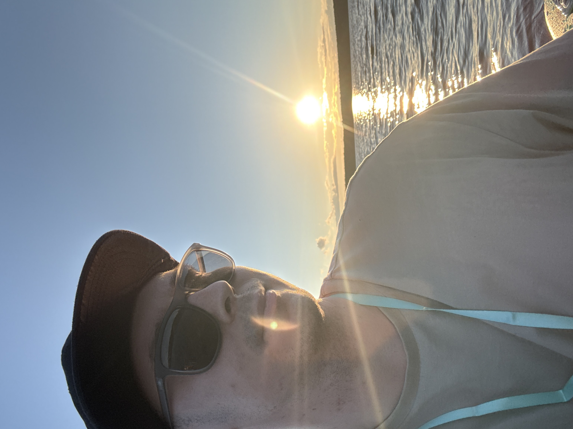 A close-up, low-angle selfie of a person wearing sunglasses and a dark baseball cap, taken outdoors. The sun is bright, low in the sky, and creating a lens flare that illuminates the scene. The person's face is angled upwards and to the left. They are wearing a light-colored shirt, possibly a tank top, with a light blue strap visible across the chest.
The background features a body of water with the sun reflecting off its surface, creating a shimmering effect. The sky is clear and blue, suggesting it is daytime. The angle of the sun and the quality of light indicate it is either sunrise or sunset, likely late afternoon or early morning. The overall atmosphere is relaxed and casual, with the bright sunlight and outdoor setting suggesting a leisurely activity, possibly near a lake or river. There is no visible text in the image. The location context provided is Polazna, Russia, which is consistent with an outdoor, possibly waterfront, setting.