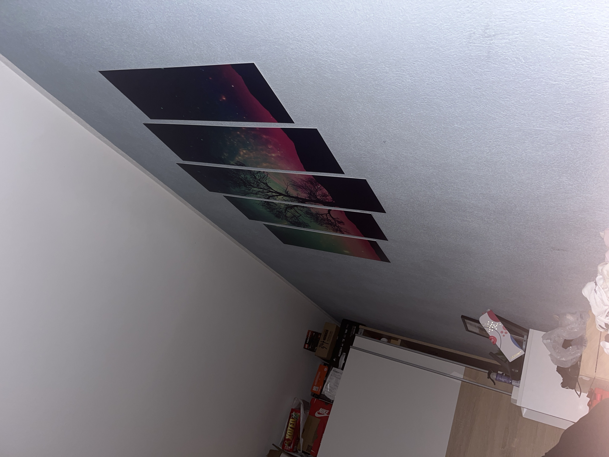 The image shows an interior view of a room. Several rectangular pieces of art with a continuous image of a tree silhouetted against a space and sky backdrop are affixed to the ceiling. The room has white walls. In the lower right, there is a white dresser with items on top of it. Adjacent to the dresser are boxes, some of which are labeled "NIKE." The environment seems to be a residential space, however, there is no mention of the location in Pokrovnik, Bulgaria. There are no people visible.