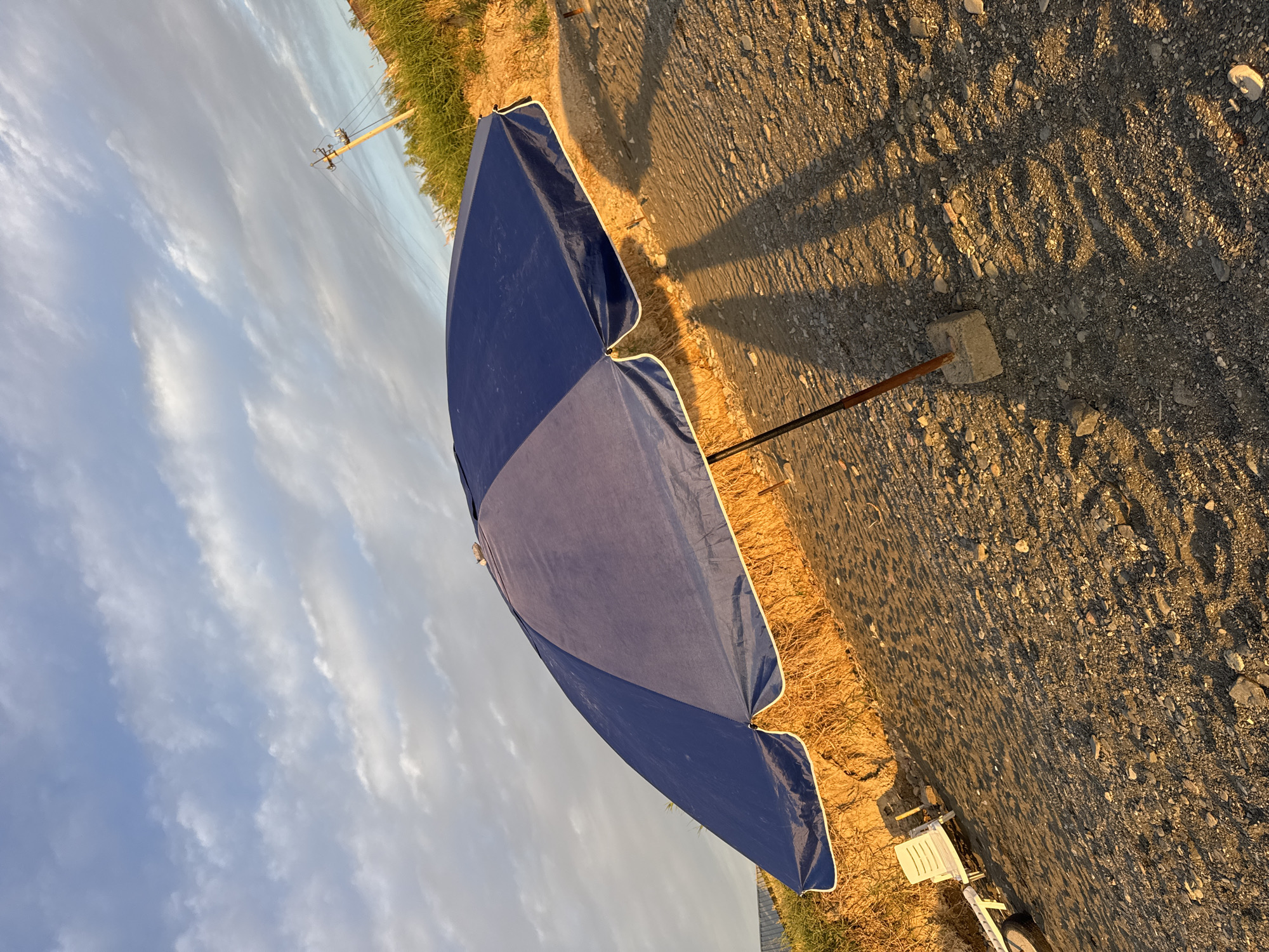 A tilted, close-up shot captures a large blue beach umbrella standing on a pebbly, sandy surface. The umbrella, with its white trim, is dominant in the frame. Behind the umbrella, dry, yellow grass grows on a slight incline, and further back, lush green vegetation is visible. A wooden utility pole with wires stretches into the distance. The sky above is filled with scattered clouds, illuminated by the warm, golden light of what appears to be either sunrise or sunset. The ground is a mix of sand and small pebbles. In the lower left corner, a white plastic lounge chair is partially visible. The overall scene suggests a quiet beach location, possibly on the outskirts of Urdzhar, Kazakhstan, bathed in the soft light of early morning or late evening.