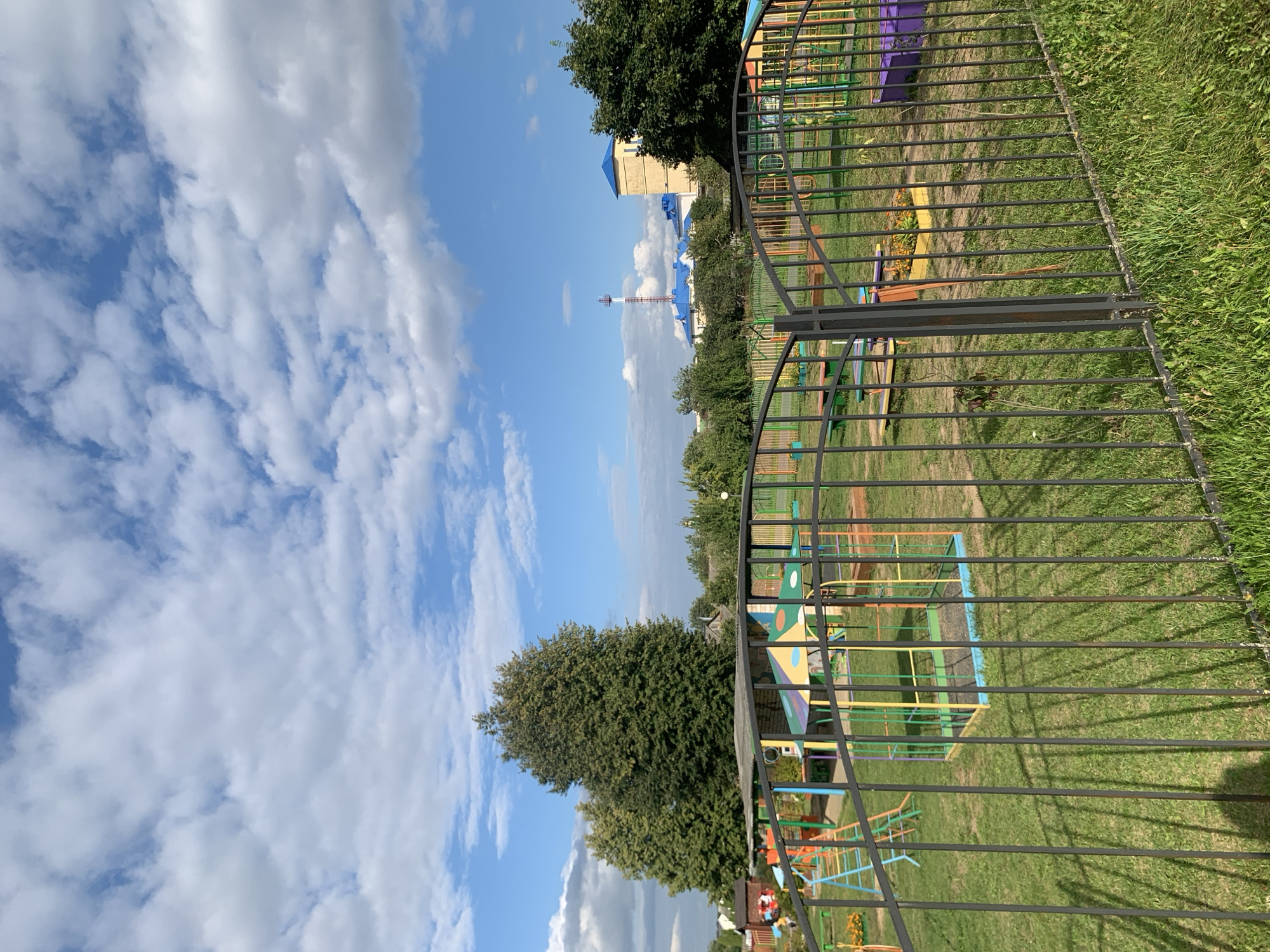 The image shows an outdoor playground in Kapyĺ, Belarus, on a sunny day with scattered clouds. In the foreground, a black metal fence separates the viewer from the playground. The playground features various colorful equipment, including slides, swings, and a sandbox. Several trees are visible in the background, along with buildings and a radio tower. The ground is covered with green grass, and there are some flowers planted in a rectangular box. The overall scene depicts a typical public park or schoolyard area.