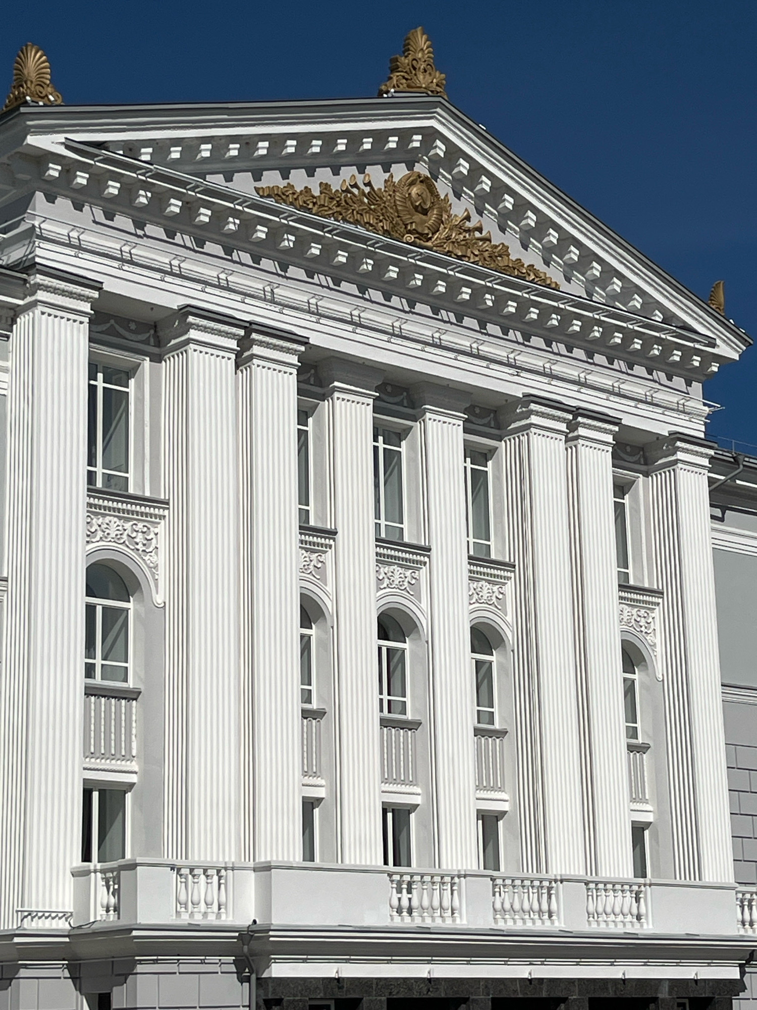 The image shows a close-up, low-angle view of the facade of the Perm State Opera and Ballet Theatre in Perm, Russia. The building features a neoclassical architectural style with prominent white Corinthian columns, ornate golden detailing, and a triangular pediment adorned with a golden emblem. Several rectangular and arched windows with white frames are visible, some with decorative elements above them. A white balcony with a balustrade runs along the facade. The sky is a clear, deep blue. No people are visible in the image.
