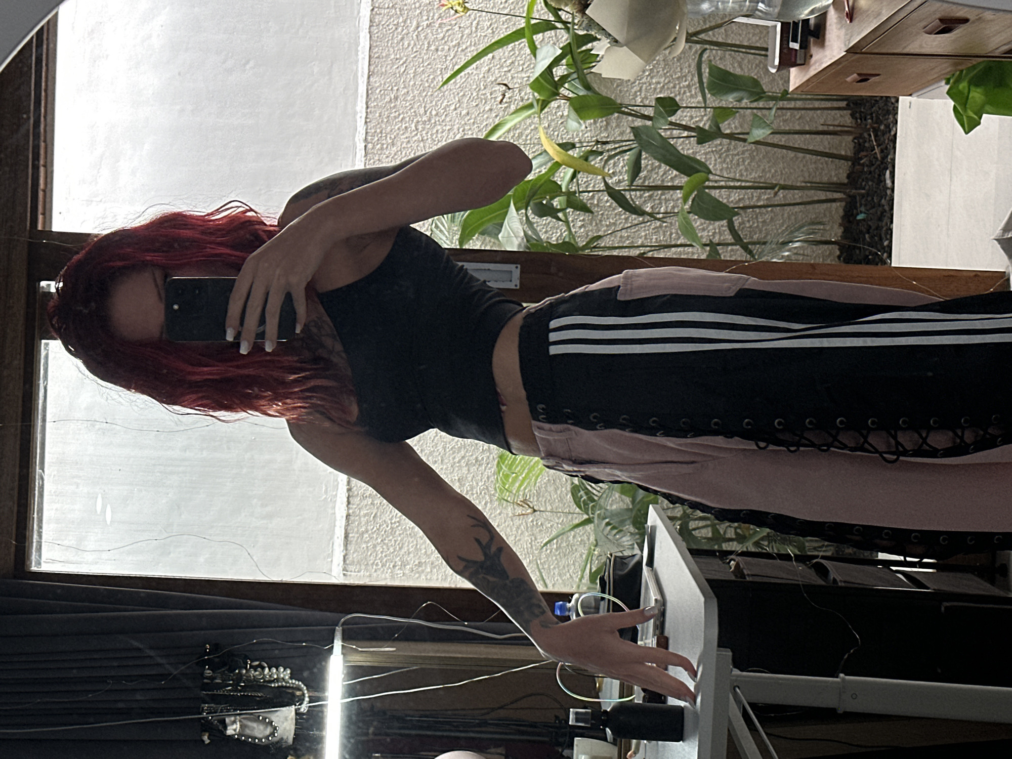 In North Kuta, Indonesia, a young woman with vibrant red hair takes a selfie in a room with large windows looking out onto a lush plant-filled area. She is wearing a black tank top and pink and black athletic pants with white stripes. Her left arm rests on a desk, and she holds a smartphone in her right hand, obscuring her face. The room appears to be a living space or studio, with various objects visible on the desk, including a bottle of spray and a notebook. The overall ambiance is casual and artistic, with a focus on natural light and greenery.