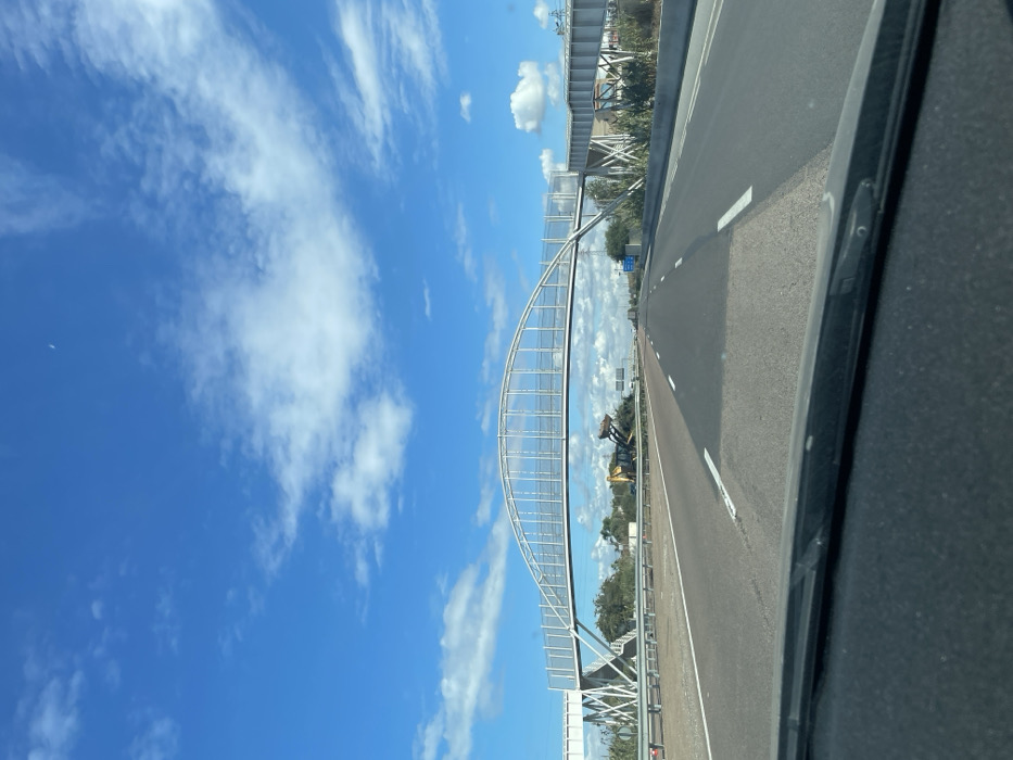 The image depicts a multi-lane highway in Skhirat, Morocco, viewed from the interior of a vehicle looking forward. A prominent white metal pedestrian overpass with an arched truss structure spans across the highway. The overpass features transparent or mesh panels along its sides and stair access on both the left and right sides of the road.
To the left of the highway's central median, beneath the overpass, a yellow articulated front-end loader or excavator is visible amidst a pile of debris or cleared vegetation. The loader's bucket is elevated. Orange traffic cones and white barriers are positioned on the median near the construction equipment.
The highway itself is asphalt, with white dashed lane markings. The pavement shows variations in tone, indicating potential repair or different material sections. The sky is bright blue with scattered white cumulus and cirrus clouds. A faint crescent moon is visible in the upper center-right portion of the sky. Sparse green vegetation, possibly shrubs or small trees, lines the shoulders of the highway. No people are visible within the frame.
