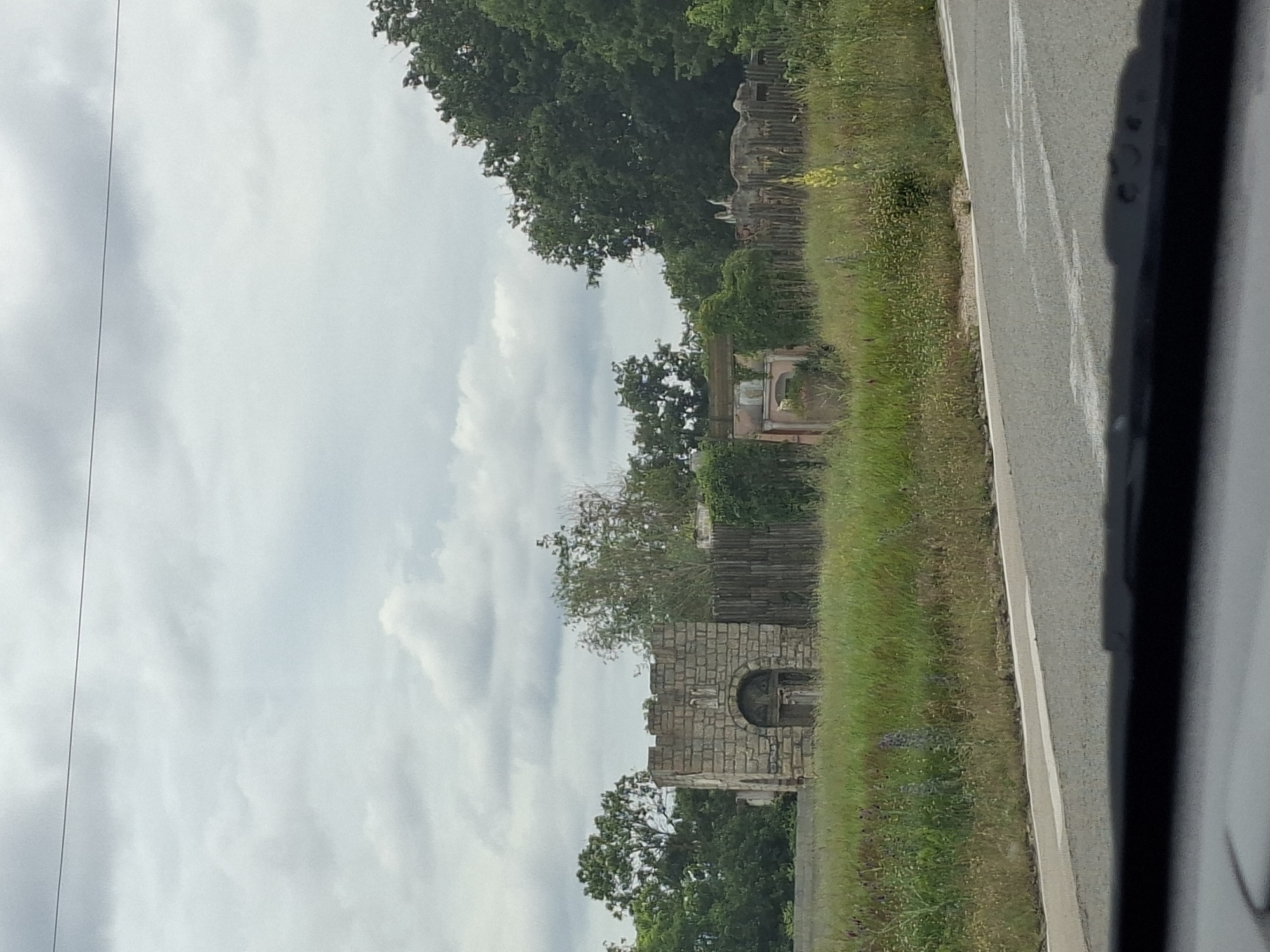 The image depicts a roadside view of what appears to be an abandoned or derelict outdoor exhibit or attraction, possibly in the vicinity of Ravna Gora, Bulgaria. In the foreground, a paved road with a white dividing line is visible, with the edge of a vehicle's dashboard or windshield in the bottom frame. Beyond the road is a field of tall, dry grass interspersed with some green vegetation and wildflowers.
In the middle ground, there are several structures. To the left, a stone-built structure resembling a small fortified gatehouse or tower stands with an arched entrance. It has a crenellated top and appears to be constructed from rough-hewn stone blocks. To the right of this structure, a weathered wooden fence runs horizontally. Beyond the fence, further to the right, are more derelict structures, including what looks like a pinkish building with arched windows and a segment of a broken wall with some decorative elements or sculptures. Scattered amongst these structures are overgrown bushes and trees. In the background, a dense line of dark green trees forms a backdrop against a cloudy, overcast sky. A thin, dark line, possibly a utility wire, stretches horizontally across the upper portion of the image. No people are visible in the scene.