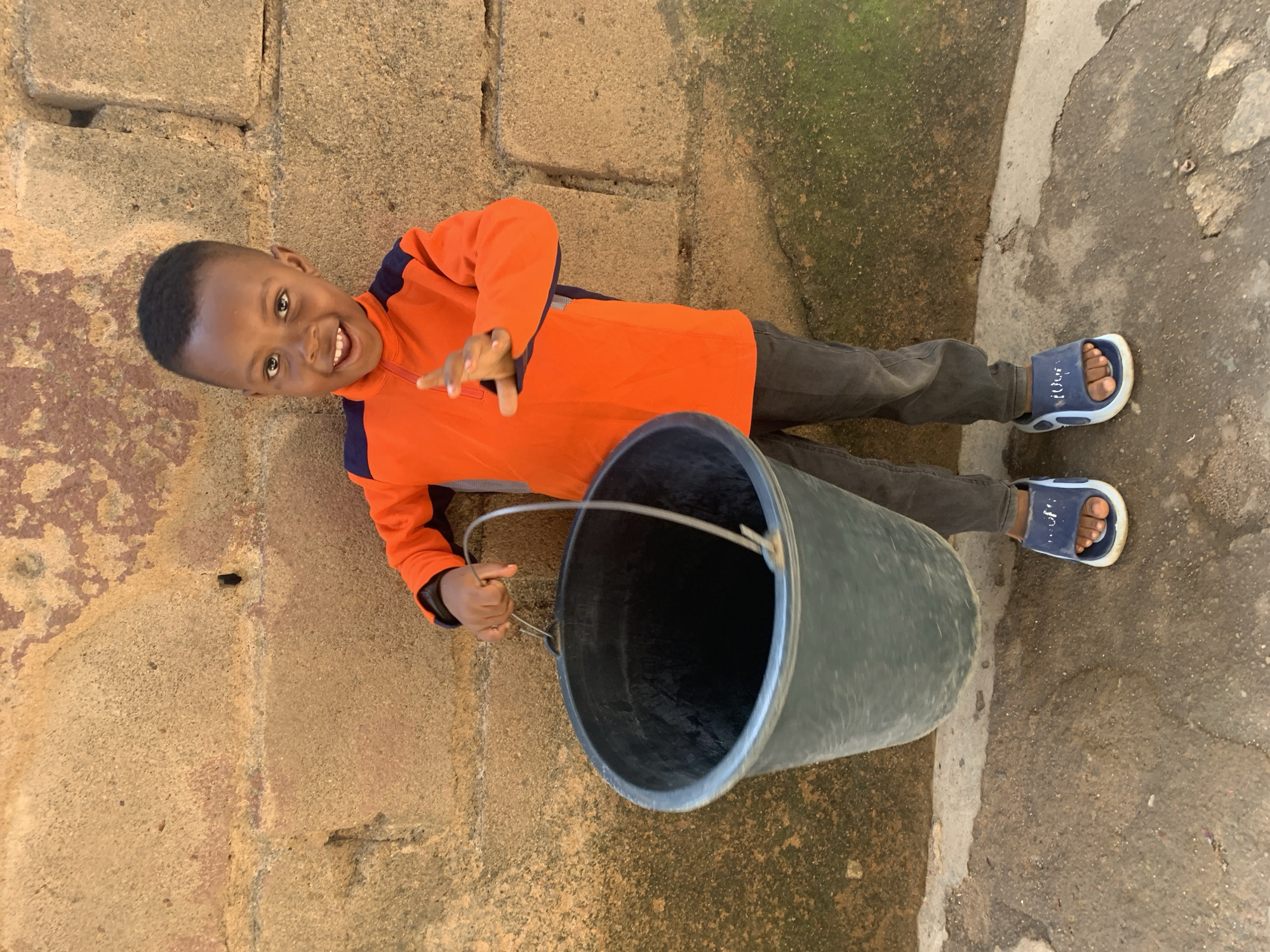 A young Black boy stands outside a building with a rough, tan-colored stone wall behind him, which appears to be in a neighborhood in Jos, Nigeria. He is wearing an orange long-sleeved shirt with dark blue accents, dark gray pants, and blue slides. The boy is holding a large, dark gray bucket by its metal handle. The bucket is empty and appears to be made of plastic. He is smiling and looking towards the camera with his left hand raised slightly. The ground in front of him is a mixture of dirt and concrete, with some moss growing on the wall.