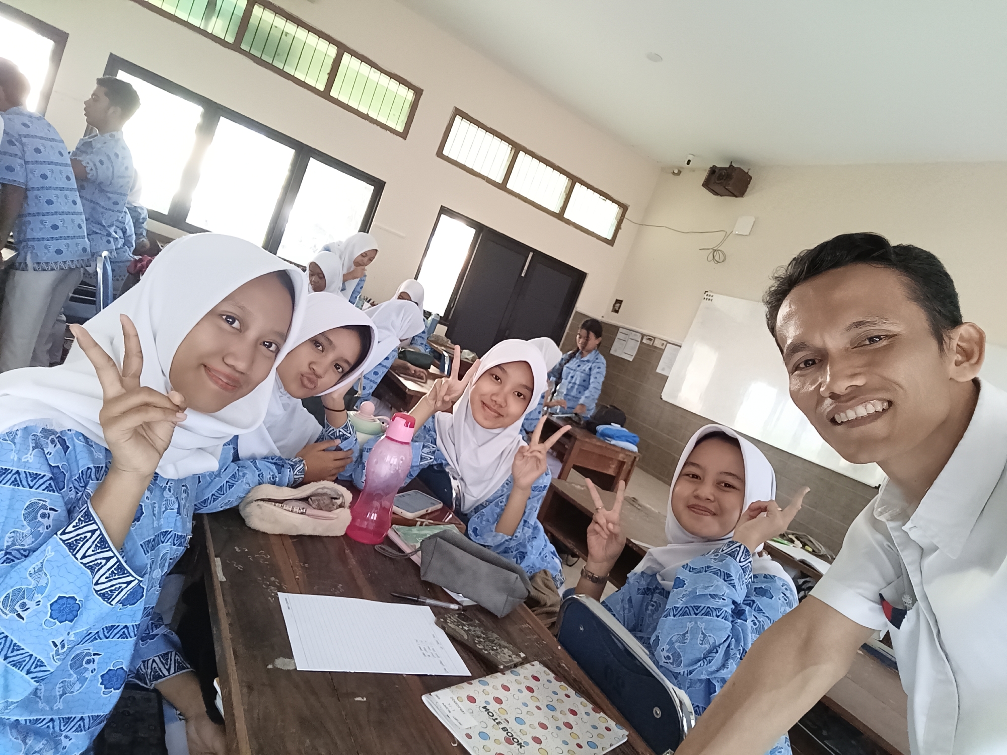 In Kediri City, Indonesia, a group of students, identifiable by their matching blue batik uniforms and white hijabs, are captured in a selfie within what appears to be a classroom setting. The students are seated at wooden desks, with one student in the foreground on the left and another on the right, both posing with peace signs. Behind them, other students are also visible, some wearing similar uniforms. A male teacher, wearing a white collared shirt, is in the foreground on the right, smiling and taking the photo. The room has large windows with green-tinted glass and a whiteboard visible in the background. Objects on the desks include notebooks, pens, and a pink water bottle. The overall scene depicts a casual and friendly interaction among students and their teacher in an educational environment.