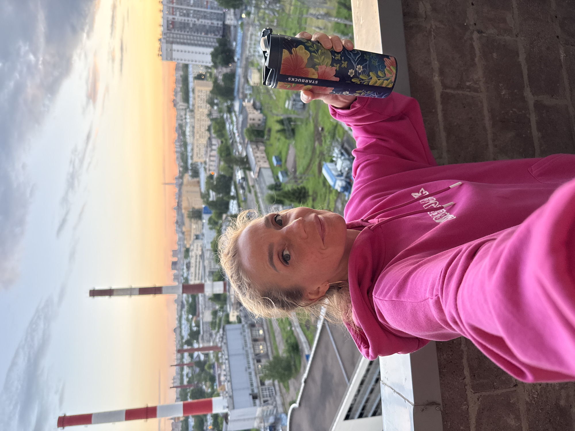 A woman with blonde hair is taking a selfie on a brick balcony with a city view. She is wearing a bright pink hooded sweatshirt and holding a dark blue travel mug with a floral pattern. The background shows industrial buildings, smokestacks with red and white stripes, and residential buildings under a sky with soft orange and grey clouds at dusk.