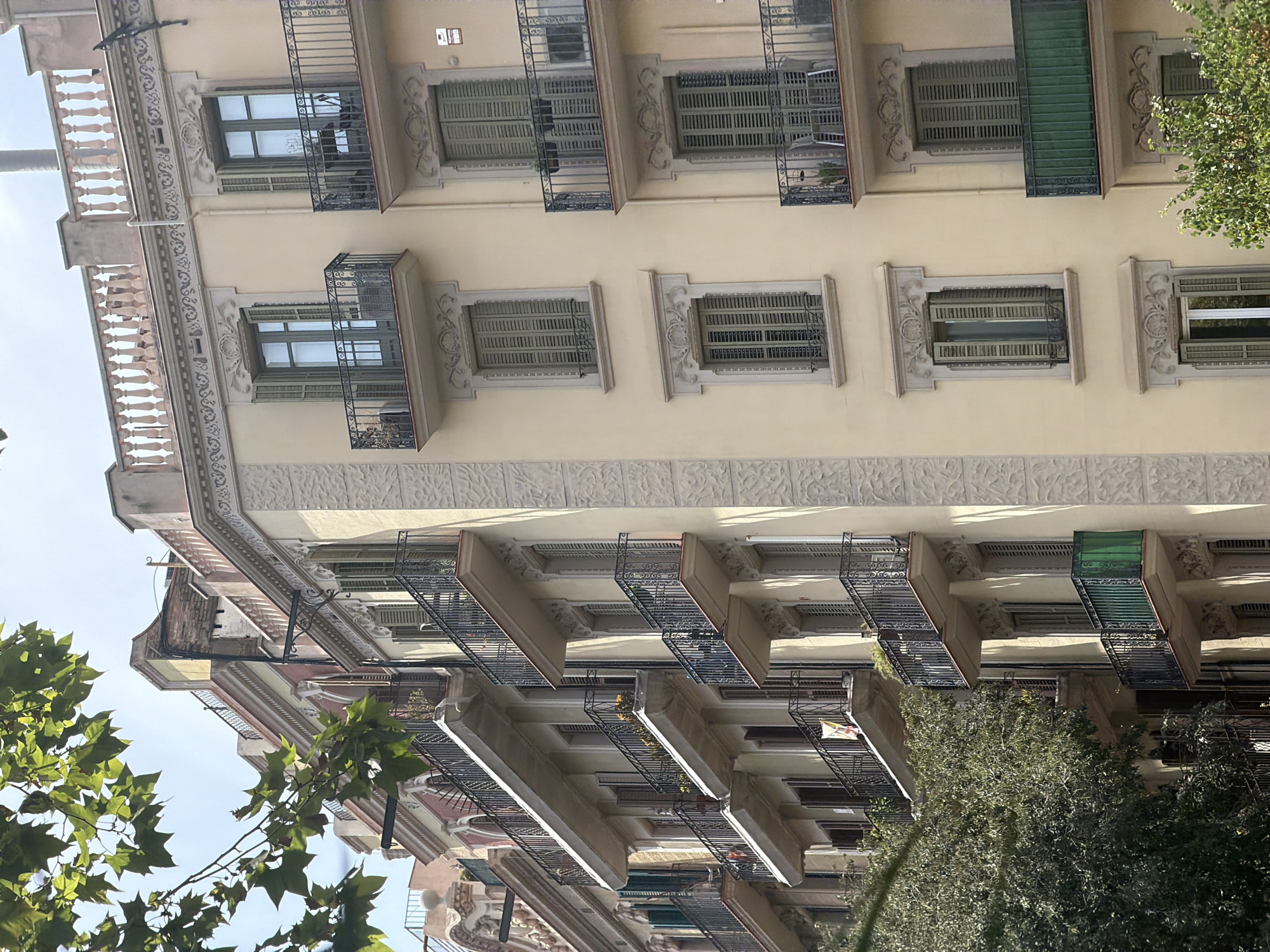 This image captures a street view of a European city, likely Barcelona, Spain, showcasing a row of apartment buildings with ornate balconies and architectural details. The buildings are several stories high, painted in light yellow and pink hues, and feature intricate moldings around windows and doorways. Numerous wrought-iron balconies with detailed patterns extend from the facades, some adorned with plants. The windows are mostly covered by green shutters, some of which are open. Green foliage from trees partially obscures the lower floors of the buildings on the left side of the frame. The sky is visible above, suggesting daytime. There are no people or discernible actions in the scene, focusing solely on the architectural elements and the surrounding environment.