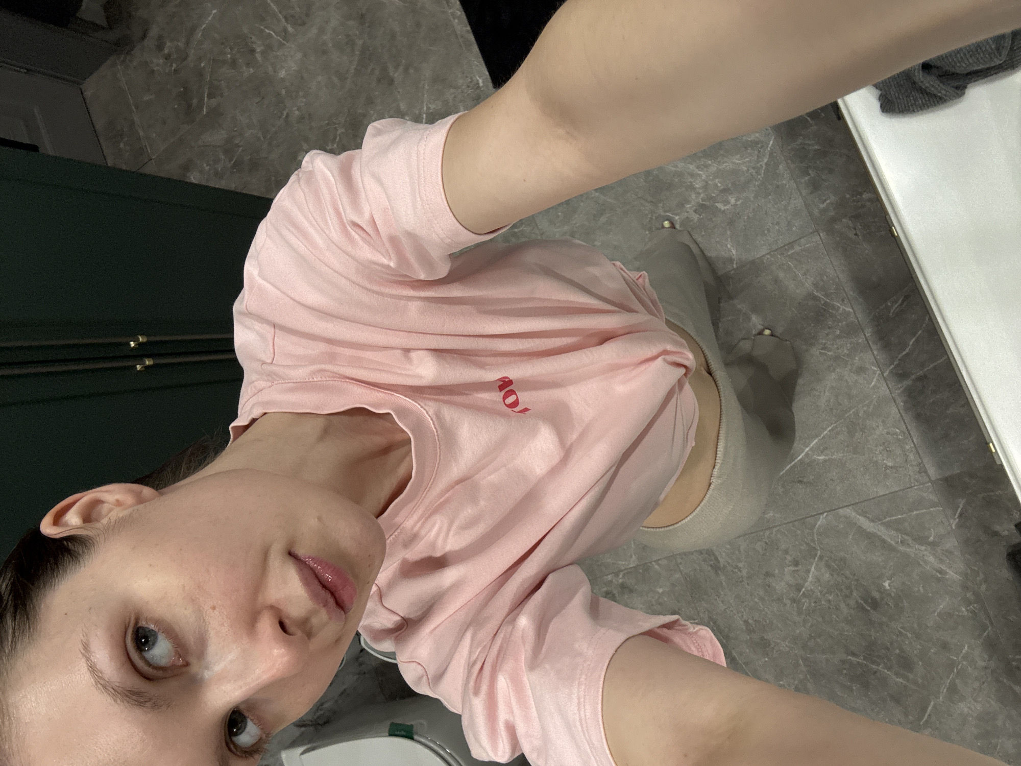 A young woman with light skin and light eyes wearing a pink t-shirt and light-colored leggings is shown in a selfie-style photograph. She is indoors, standing on a gray tiled floor. In the background, there is a green cabinet or wardrobe and a white counter or sink. The location cannot be determined from the image, so information about the city of Tomilino, Russia, is not applicable.