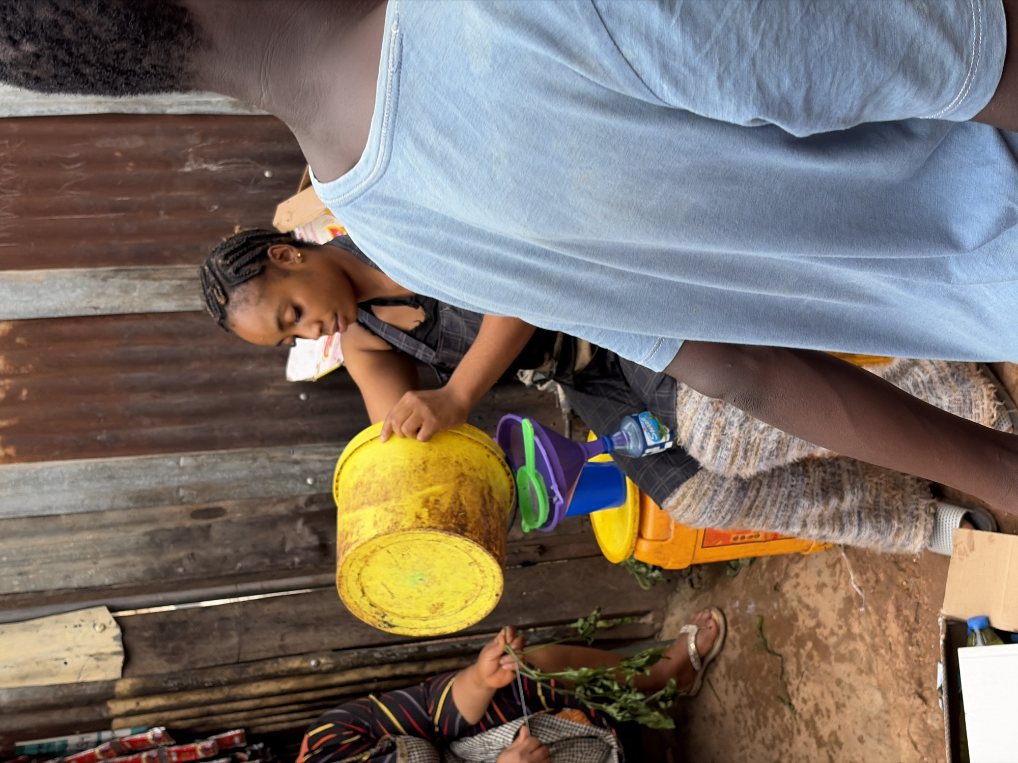 The image shows a woman with braided hair, wearing a black top with overalls, working with a yellow bucket and a stack of colorful funnels. She appears to be pouring or transferring something. Another person is seated nearby, handling green leafy plants, possibly herbs or vegetables. The location appears to be outdoors, with a backdrop of wooden or corrugated metal walls. The ground is earthen. The photo was taken in the city of Jos, Nigeria. Another person is standing above the woman with the bucket, with their back to the camera, wearing a light blue shirt.