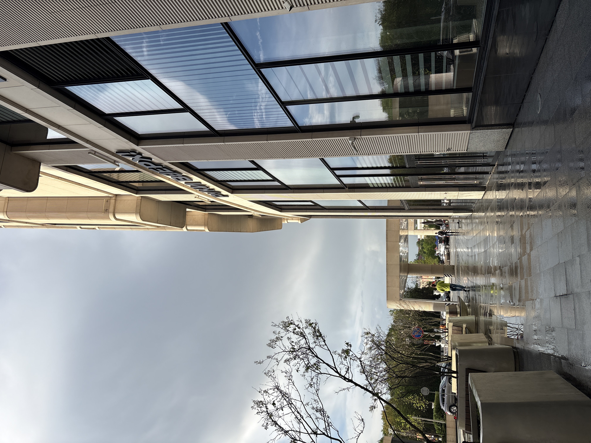 The image presents an outdoor urban scene, likely in Minsk, Belarus. The camera seems to be angled upwards towards modern architecture. The building features a glass facade reflecting the sky and nearby trees. A sign is visible on the building. The ground is paved with what appears to be wet stone, reflecting the surroundings. A bare tree stands prominently in the foreground. In the background, people are seen walking. There are also planter boxes. The sky is overcast, suggesting recent rain.