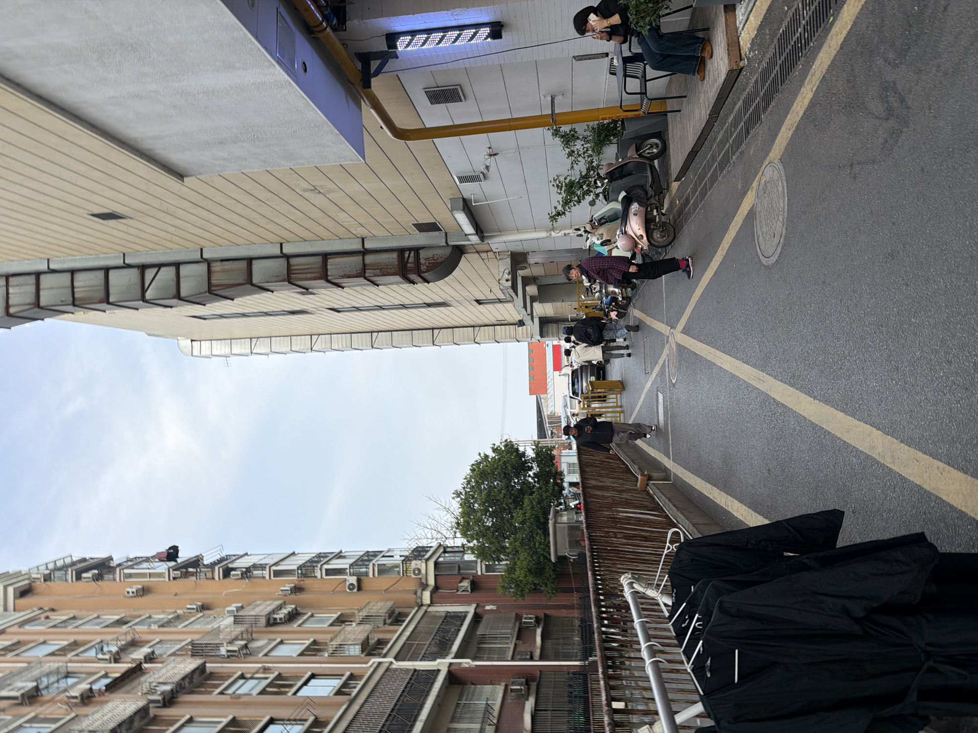 The image shows a street scene in Jiangning District, China. On one side of the street is a multi-story building with many windows, some with air conditioning units. Clothes are hung on a rack outside one of the lower floors. On the other side of the street, there is a mix of buildings, including a taller structure with horizontal siding. The street is paved and has yellow lines indicating parking or lane divisions. Several scooters and people are present on the street and sidewalk on the right side of the image. One person is seated, talking on a phone. The sky is visible above the buildings.