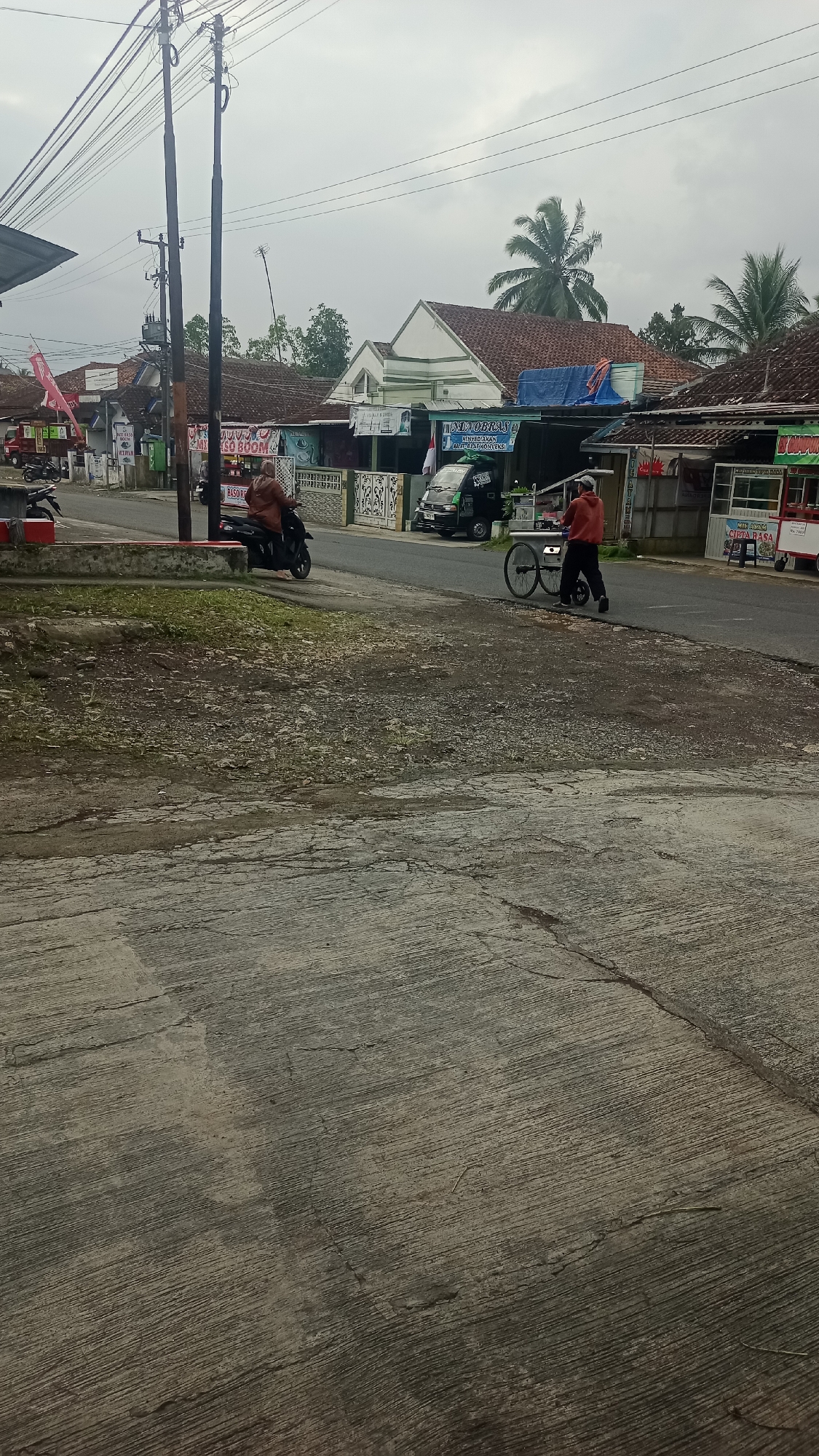 The image depicts a street scene in Karyamandala, Indonesia, with a focus on a road and sidewalk area. In the foreground, a textured concrete surface with visible cracks and debris occupies the lower portion of the frame. Beyond this, a dirt and gravel area with sparse green vegetation leads to the road.
On the road, a person wearing a brown hijab and jacket rides a black motorcycle, heading away from the camera. To the right of the motorcycle, a person in an orange hoodie and black pants pushes a wheeled cart, also moving away from the camera.
The background reveals a row of buildings with tiled roofs, characteristic of Indonesian architecture. Several signs are visible on these buildings, some with Indonesian text. A dark green van is parked on the side of the road. Electrical poles with numerous wires are prominent in the mid-ground, suggesting a developed, albeit somewhat rustic, urban or semi-urban environment. The sky is overcast, indicating a cloudy day. The overall impression is one of a typical street in a residential or commercial area within Karyamandala.