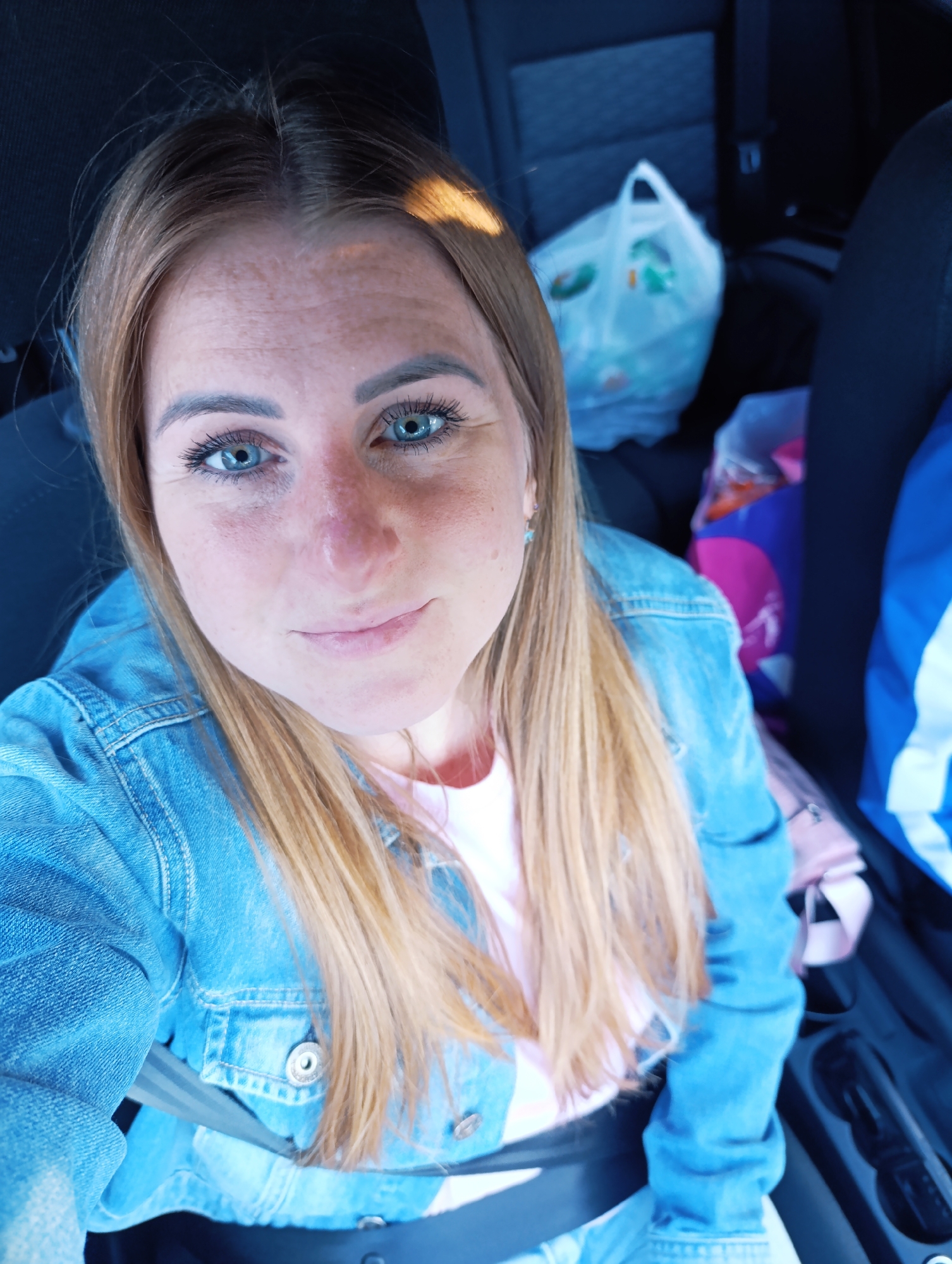 This media file is a selfie of a young Caucasian woman with long, blonde hair and bright blue eyes, taken from a high-angle perspective inside a vehicle. She is wearing a denim jacket over a pink t-shirt. Her expression is a slight smile, and she appears relaxed. Her eyes are looking directly at the camera. The lighting suggests it is daytime, possibly late afternoon, as there is a warm light casting shadows on her forehead.
The background shows the interior of a car, with a black headrest visible behind her. To her right, there are several shopping bags, one of which is a white plastic bag with some green markings, and another is a purple bag with a pink handle, likely indicating she has been shopping or is on a trip. A blue and white striped object, possibly a bag or part of a vehicle seat, is also visible in the far right of the frame. The seatbelt across her chest is clearly visible, indicating she is seated in the driver's or passenger's seat.
The scene is an indoor setting within a car, with elements suggesting a casual outing or travel. No text is visible in the image. The overall mood is informal and personal.