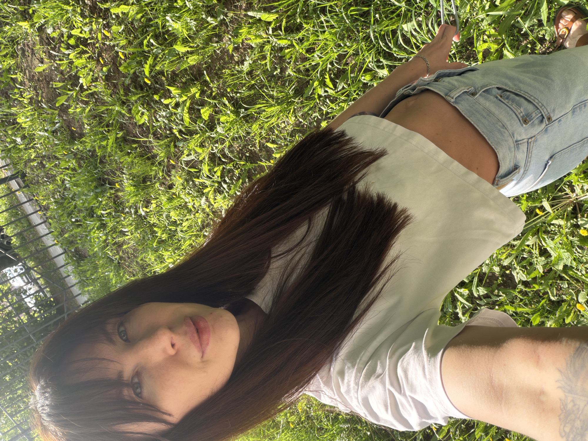 A fair-skinned female lies supine on grass in an outdoor setting. She has long brown hair, light blue eyes, and is wearing a white short-sleeve shirt and light blue jeans. The location appears to be in Perm, Russia, indicated by the presence of green grass and trees. The woman is holding a phone, possibly taking a selfie. A metal fence is visible on the left side of the image. Her left arm, which has a tattoo, is extended.