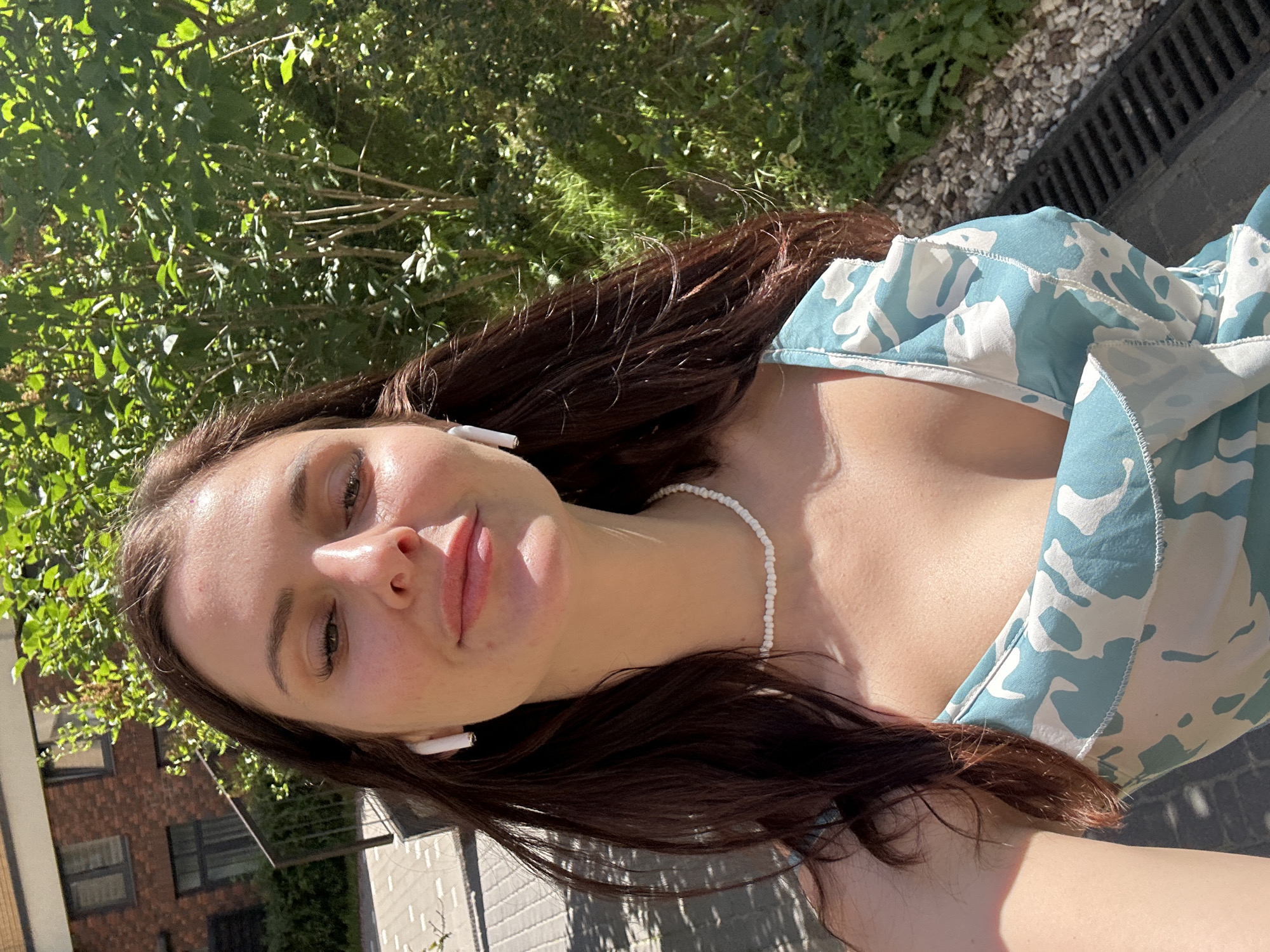 A young woman with long brown hair, wearing white wireless earbuds and a light blue and white patterned dress, stands outdoors in front of lush green foliage and a brick building. The woman is looking directly at the camera, with her lips slightly parted. She is also wearing a white beaded necklace. The background includes a pathway with stone pavers and a dark metal grate. The lighting suggests it is daytime with sunlight casting shadows.