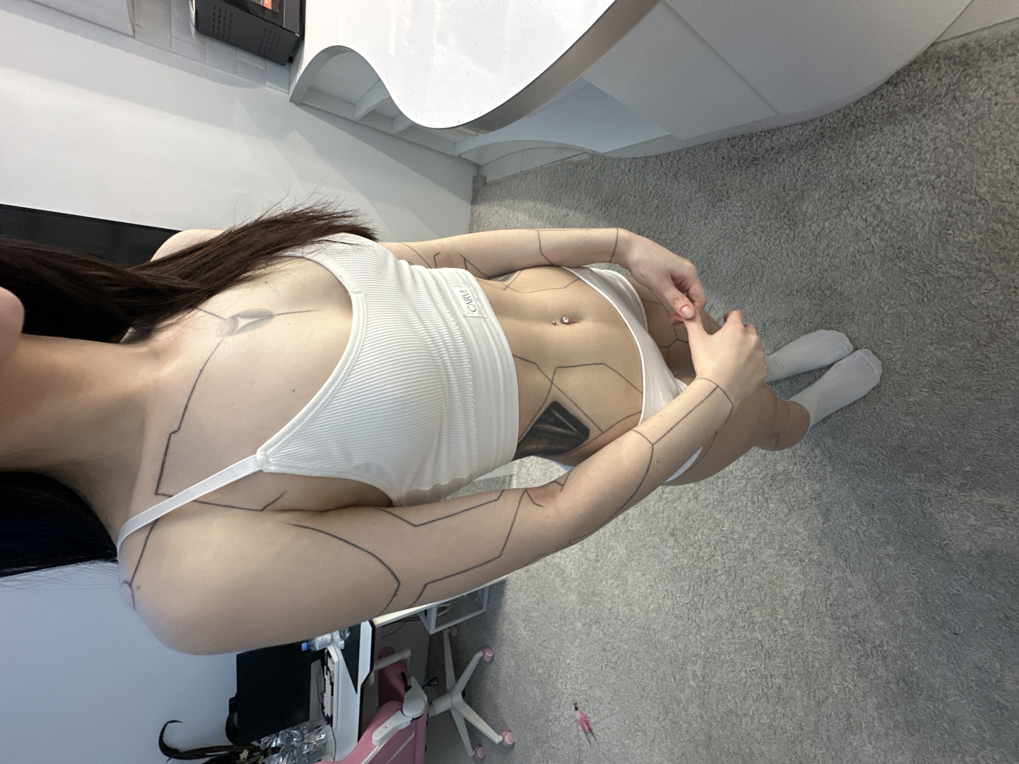 A woman with dark hair is lying on a gray carpet, oriented horizontally in the frame. She is wearing a white ribbed tank top and white underwear. She has a navel piercing and geometric line tattoos on her skin. A white desk or counter is partially visible in the upper center and left of the image. A black object is mounted on the wall in the upper left corner. A white desk and pink and white chair are partially visible in the lower left corner. The background shows a white wall and a gray carpeted floor. There is no information available about the location being in the city of Khimki, Russia.