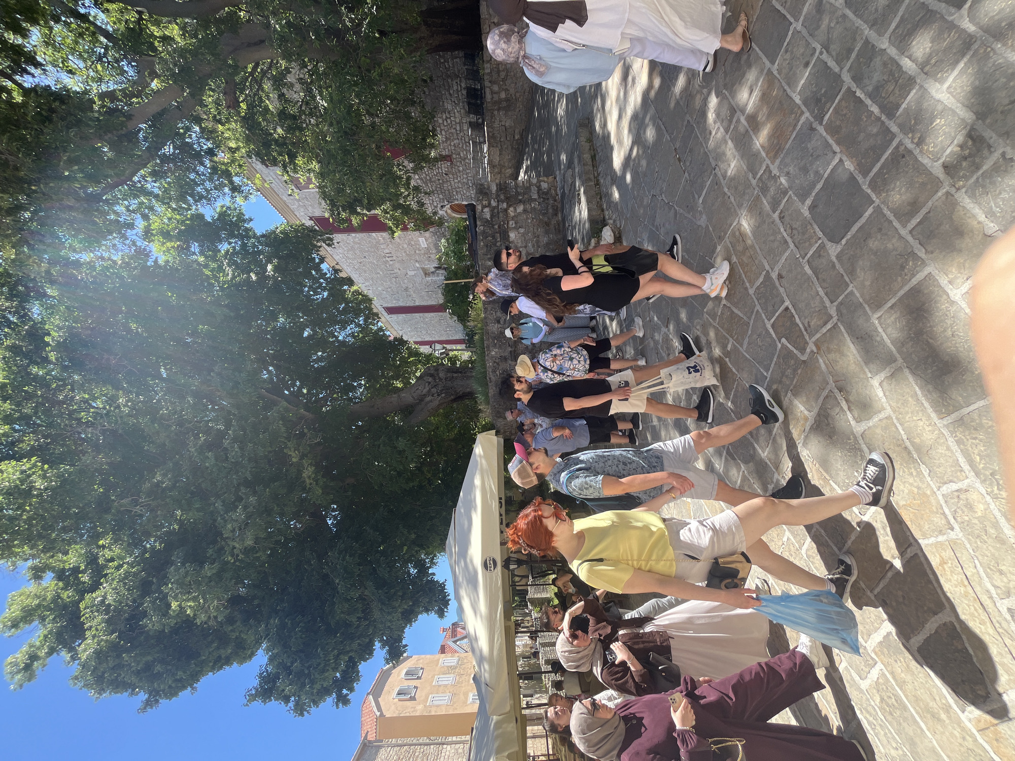 The image captures a group of people walking along a paved pathway in Budva, Montenegro. The pathway is constructed with large, rectangular stone blocks. A large tree with dense green foliage provides shade over part of the pathway. To the left, a building with a red-tiled roof and beige facade is visible. On the right, parts of a stone building are visible. The people in the group are dressed in casual summer clothing. A Pepsi branded awning is visible in the foreground. The sky is clear and blue.