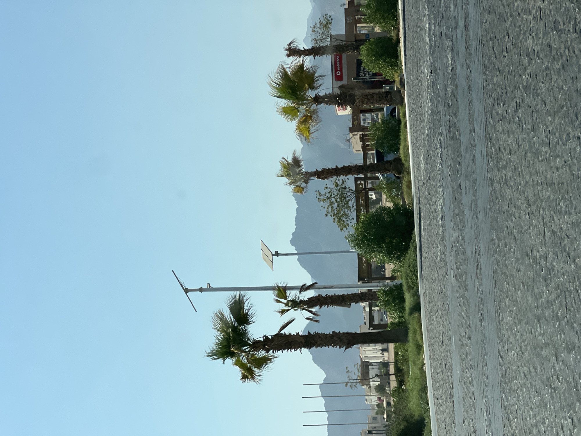 In Sharm El Sheikh, Egypt, a paved road made of grey cobblestones occupies the foreground of the image. Beyond the road, a line of palm trees stands tall, with some appearing to be in various states of health. Behind the palm trees, a low-rise building with white facades is visible, suggesting a commercial or residential area. A sign with the Vodafone logo is also present on one of the buildings. In the background, rugged mountains create a dramatic silhouette against a clear blue sky. A solar-powered street light is also visible. The scene appears to be outdoors, under bright sunlight, and is characterized by a dry, arid climate.