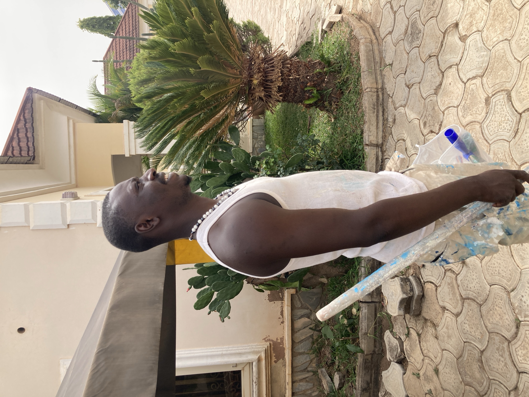 A Black man is depicted in an outdoor residential setting, looking upwards with a joyful or smiling expression. He wears a white ribbed tank top and a white and black beaded necklace. His head is tilted back, and his gaze is directed towards the sky. He has very short, dark hair.
He is holding a long tool, likely a paint roller, in his right hand. The roller is partially covered in a plastic bag or wrap and shows splatters of blue paint. Some blue plastic items, possibly bottles or caps, are visible inside the plastic covering. This suggests he may be engaged in painting or renovation work.
The ground beneath him is covered with distinctive light-colored, interlocking concrete pavers, with grass growing in the spaces between them and along the edges. In the mid-ground, a large sago palm tree stands prominently, surrounded by other green foliage including several prickly pear cacti with flat pads and some small reddish fruit or buds.
In the background, a house with cream-colored walls and reddish-brown tiled roofs is visible. The architecture features decorative elements and cornices. The sky is bright and appears light gray or white, indicating a daytime setting. The overall environment suggests a residential area, consistent with the location context of Jos, Nigeria. There is no visible text in the image.