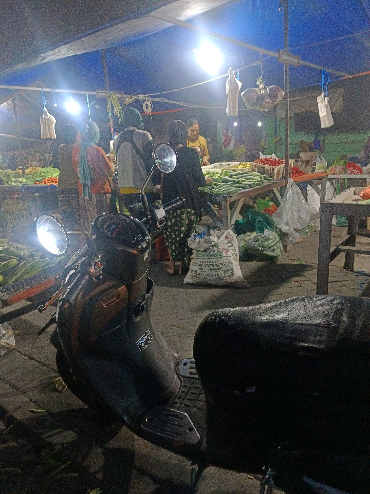 The image depicts a nighttime scene at a market in Wonocolo, Indonesia. In the foreground, a dark-colored scooter is parked, with its headlights on, casting bright illumination. The scooter's seat appears worn. Behind the scooter, a market stall is visible, covered by a blue tarp supported by wooden poles. Bright lights hang from the tarp, illuminating the area.
Several individuals are present at the market. Three people are standing near a stall piled with vegetables, including cucumbers and tomatoes. Another person, wearing a yellow shirt, appears to be working at a stall behind a display of red chilies. Several bags, some containing produce, are scattered around the market. A large, white sack with Indonesian writing is prominent on the ground. The ground is paved.
