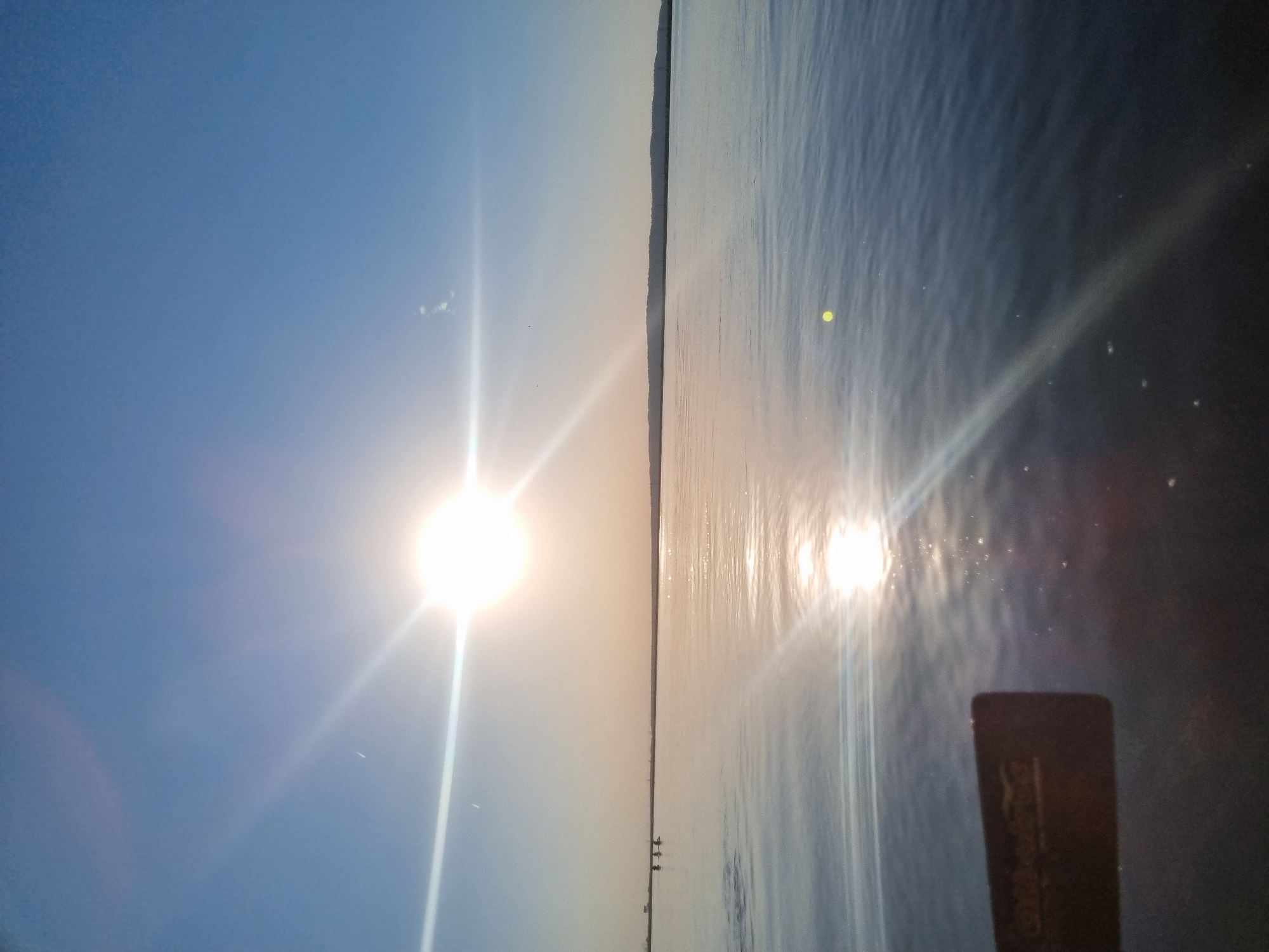 The image shows a body of water bisected by a thin strip of land, likely a distant shore. On the left side of the image, there is a bright sun in a clear blue sky. The sun's reflection is visible on the water on the right side of the image. In the bottom right corner, there is an object which partially obscures the view. Given the location (Chaikovsky, Russia), this is possibly a view of the Kama River.