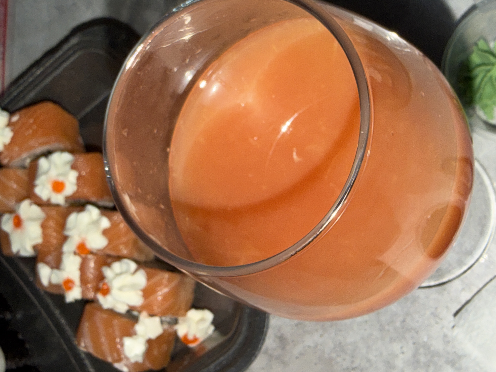 An overhead shot captures a glass filled with an orange-colored beverage, likely juice or a smoothie, prominently positioned on a light-colored surface. The glass is tilted, and reflections are visible on its clear surface. To the left and partially obscured by the glass, several sushi rolls are arranged in a dark, rectangular tray. These rolls are wrapped in salmon and topped with dollops of white cream cheese or a similar sauce, each adorned with a small, bright orange garnish, possibly masago or another condiment. In the upper right background, a small, clear container holding a green substance, likely wasabi, is visible. The overall scene suggests a casual meal or snack setting. No people are visible, and no text is discernible. The time of day and weather cannot be determined from the indoor setting. The location context for this image is Chaikovsky, Russia.