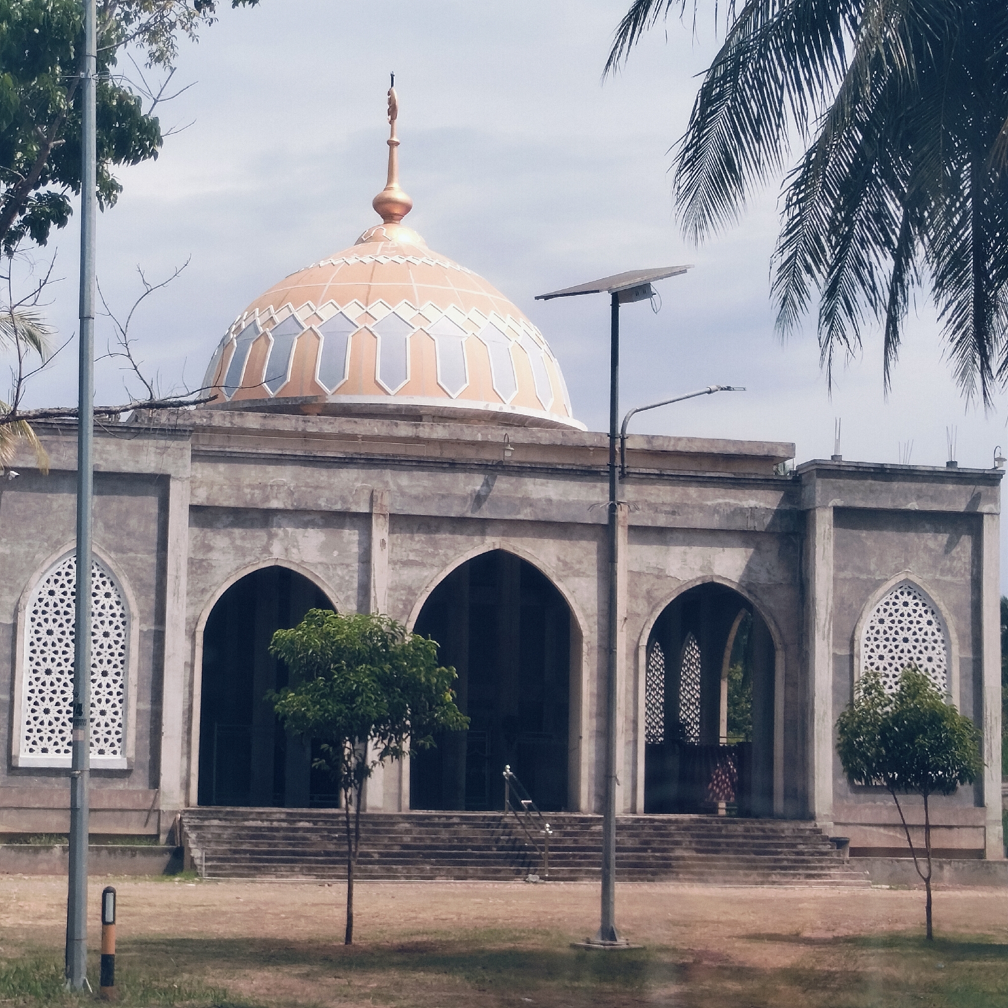 The image displays a mosque in Cottupah, Indonesia, under a bright, overcast sky. The central structure features a large, ornate dome, predominantly orange-gold with intricate grey and white geometric patterns, topped by a golden finial. The building's facade is constructed from grey concrete, presenting a series of symmetrical arched openings and two distinct windows with elaborate white geometric lattice designs on either side. Concrete steps lead up to the mosque's entrances from a grassy-dirt foreground.
Two utility poles are visible; one on the far left, partially obscured by a tree, and another on the right featuring a solar panel and a light fixture. Green foliage dots the scene, including a larger tree on the left, several smaller trees in front of the mosque, and a large palm frond arching into the frame from the right. A faint figure in patterned clothing can be discerned within one of the arched doorways on the right side of the building.
The scene conveys a sense of calm and stillness, indicative of daytime, possibly late morning or early afternoon, given the diffused light. No specific activity or event is taking place, and no visible text appears in the image. The architectural style, including the dome and arched elements, is characteristic of Islamic design.