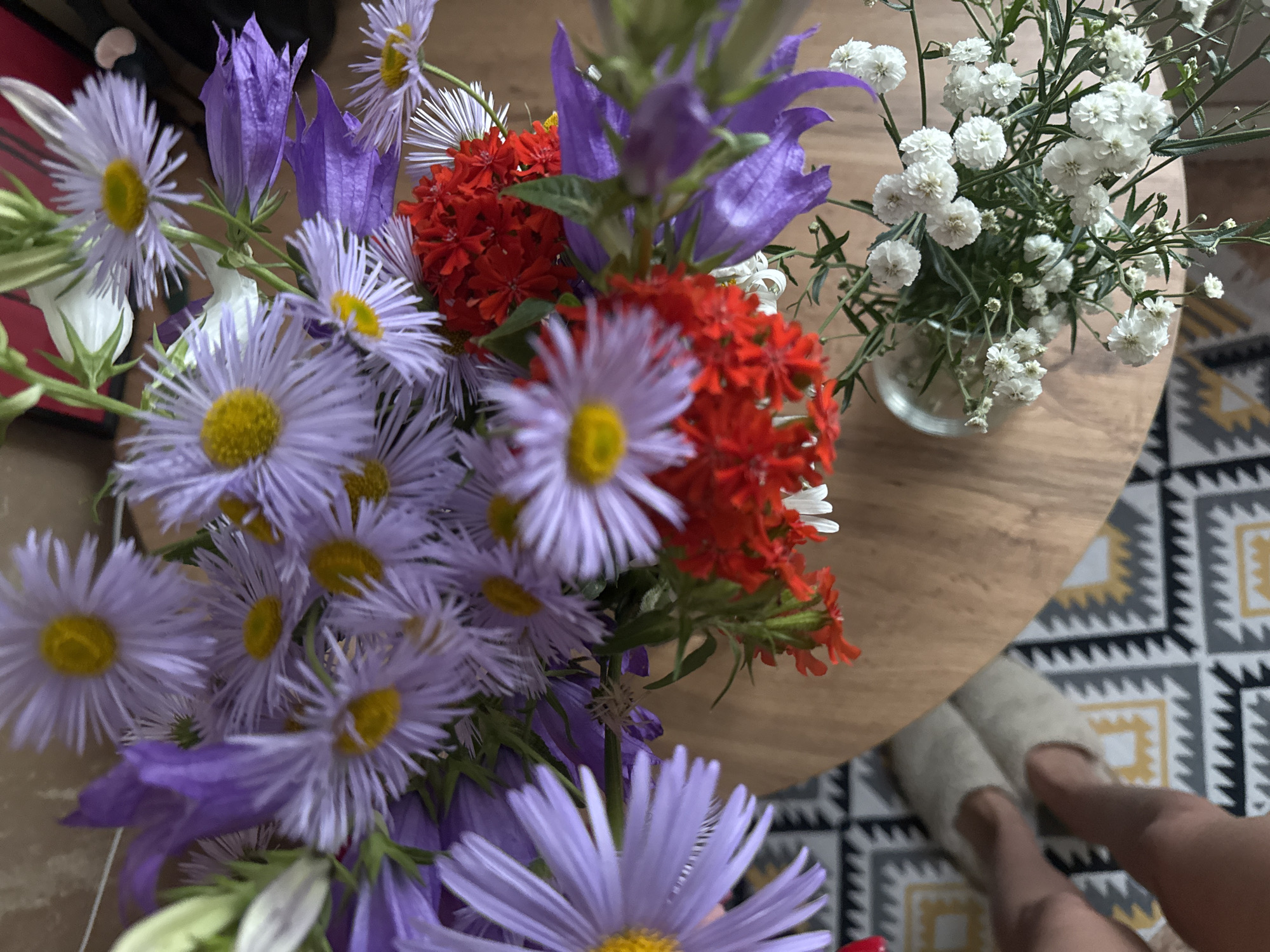 A close-up, top-down view captures a vibrant bouquet of flowers arranged on a wooden surface. The bouquet features an assortment of purple asters with yellow centers, striking red clusters of small flowers, and delicate white blooms interspersed with greenery. Among the flowers, purple bell-shaped blossoms are also visible. The arrangement is presented in a clear glass vase. To the left of the vase, a person's feet, clad in light-colored slippers, are partially visible. The scene is set against a patterned rug with a geometric design of black, white, and yellow zigzags. The photograph was taken indoors, with no specific indication of the city of Chaikovsky, Russia, beyond the prompt's request.