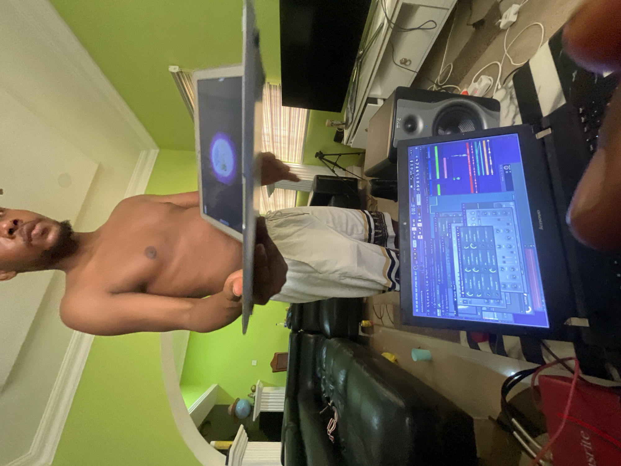 A shirtless man stands in a room in Abuja, Nigeria, holding one laptop while another rests on a table. Both laptops display visual content. A black leather couch is nearby, and audio speakers are visible on a shelf. The room is painted green.