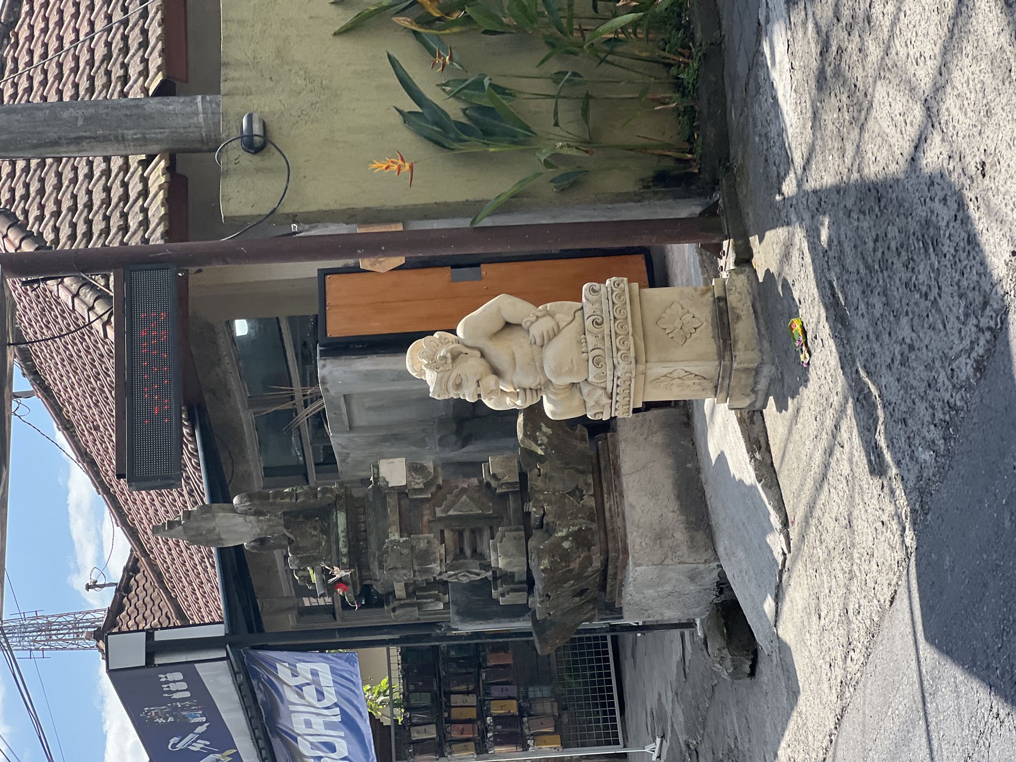 A statue of a stout, bald, human-like figure sits on a pedestal in front of a building in Ubud, Indonesia. The figure is depicted with its knees pulled up towards its chest, and its hands resting on its legs. It has a round face with a prominent nose and mouth. The statue is made of stone and appears aged. To the left of the statue is a larger, more elaborate stone structure, possibly an altar or shrine, with intricate carvings. Behind these structures, a building with a red-tiled roof is visible, along with a blue banner that reads "SORIES". A tall radio tower can be seen in the background. The ground is paved with rough concrete. The scene is lit by bright sunlight, casting distinct shadows.