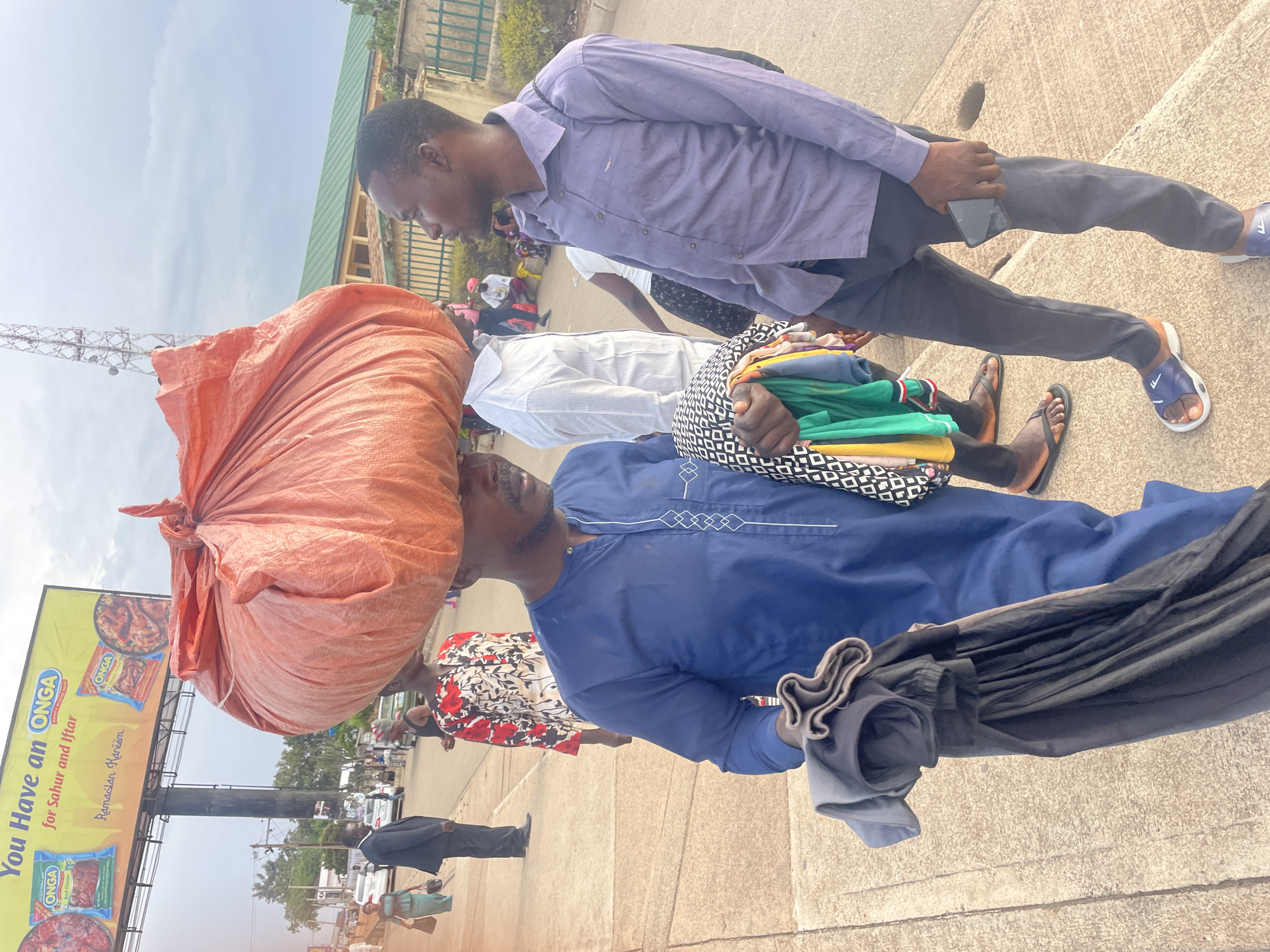 This image, presented at a 90-degree counter-clockwise rotation, depicts a bustling street scene in Jos, Nigeria, under clear daylight conditions.
In the immediate foreground, a man wearing a blue traditional kaftan with white embroidery on the chest carries a substantial orange bundle, possibly containing goods or textiles, balanced on his head. He also holds a collection of neatly folded garments in shades of green, yellow, black, and grey in his left arm. Adjacent to him, another man dressed in a long-sleeved purple shirt and dark trousers stands, holding a smartphone in his right hand. He is wearing blue and white flip-flops. Both men appear to be engaged in a moment of pause or interaction.
Behind these two individuals, other people are visible, including someone in a white striped shirt and a woman partially seen wearing a floral dress. In the background, further down the street, more pedestrians are present, with one man distinctly visible in a black suit.
To the left side of the frame, a prominent yellow billboard stands on a metal structure. The billboard advertises "ONGA" seasoning, with text reading: "You Have an ONGA for Sahur and Iftar. Ramadan Kareem." The ad features product packaging and images of food. A tall cellular tower or mast is visible against a bright, clear sky above and to the left of the billboard. Portions of buildings, including one with a green roof, are visible in the background, suggesting an urban or semi-urban setting. The ground beneath the people is paved, likely concrete or asphalt. The scene portrays typical daytime activity in a busy public space.