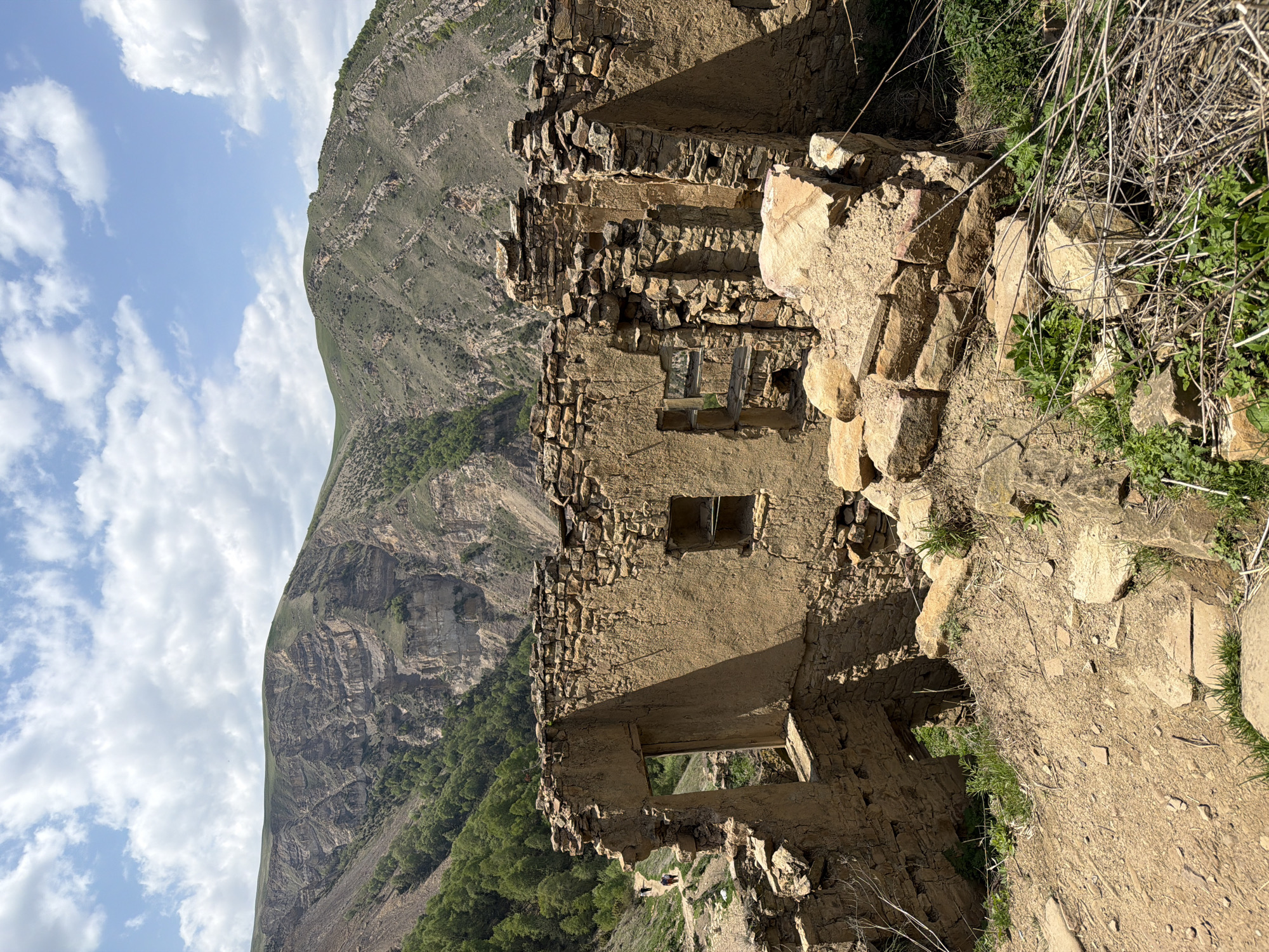 The image shows the ruins of a stone building in a mountainous environment, possibly located in the city of Unknown, Unknown. The building is severely damaged, with crumbling walls and missing sections. Some small windows are visible in the remaining walls. The surrounding environment includes a large mountain slope with visible geological layers and some vegetation. The sky is partly cloudy. The foreground is dominated by a rocky, earthy terrain. There are no people visible in the image.