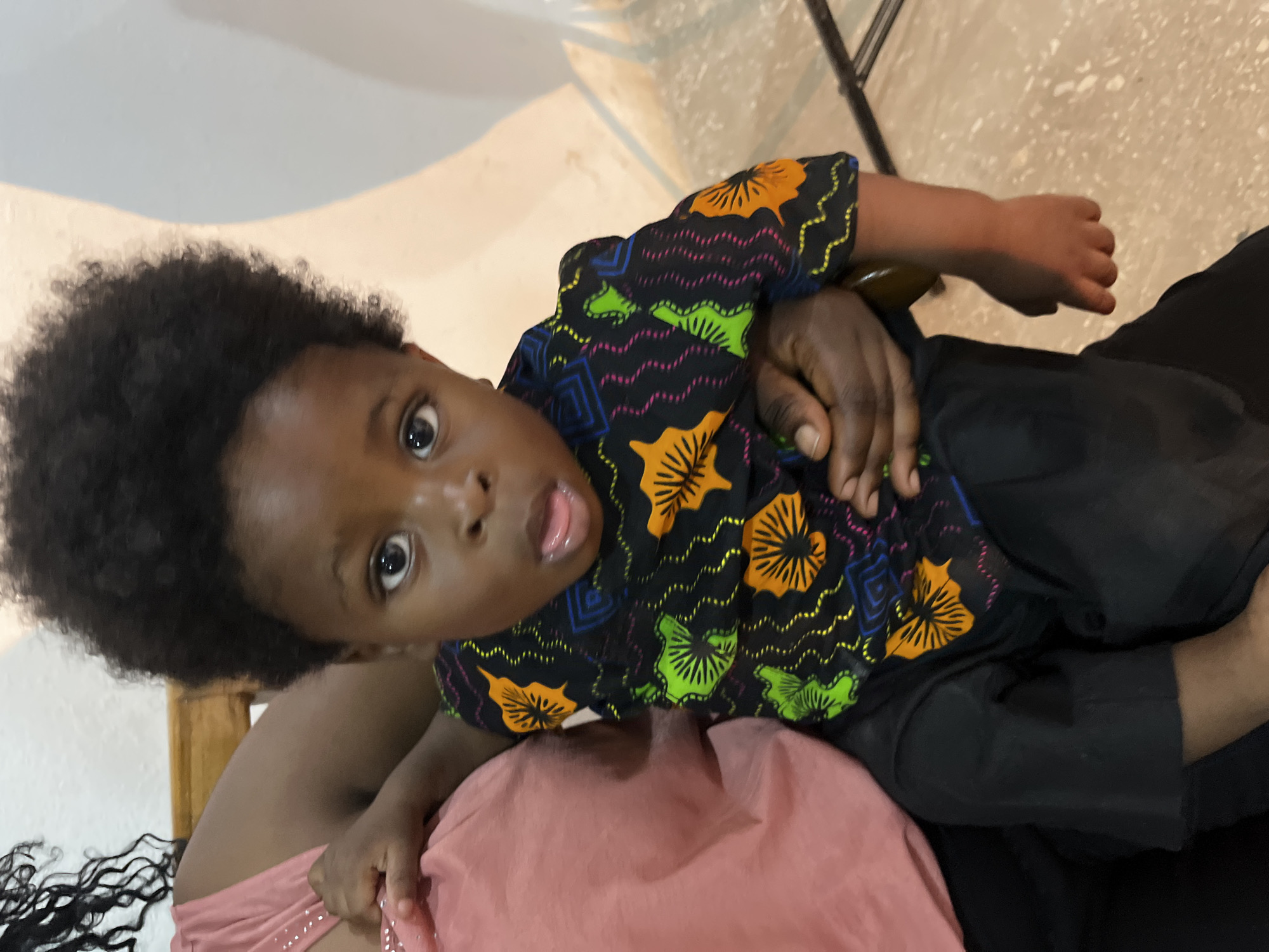 This media file shows a close-up, high-angle shot of a young Black child being held. The child has dark, curly hair and is looking directly at the camera with wide eyes and an open mouth, appearing surprised or excited. The child is wearing a black shirt with a colorful, abstract pattern featuring orange, green, and blue shapes, possibly representing flowers and geometric designs. The child's left arm is visible, held close to their body.
The child is being held by an adult, whose arms and torso are partially visible. The adult is wearing a pink garment, likely a dress or top, and black pants. The adult's hands are visible, one holding the child's arm and another resting on their lap. The background is blurred and appears to be an indoor setting with light-colored walls and a tiled floor. A wooden chair is partially visible behind the child. The lighting suggests it could be daytime, but the exact time is not discernible. The overall mood conveyed is one of warmth and intimacy, focusing on the child's expression. There is no visible text in the image.