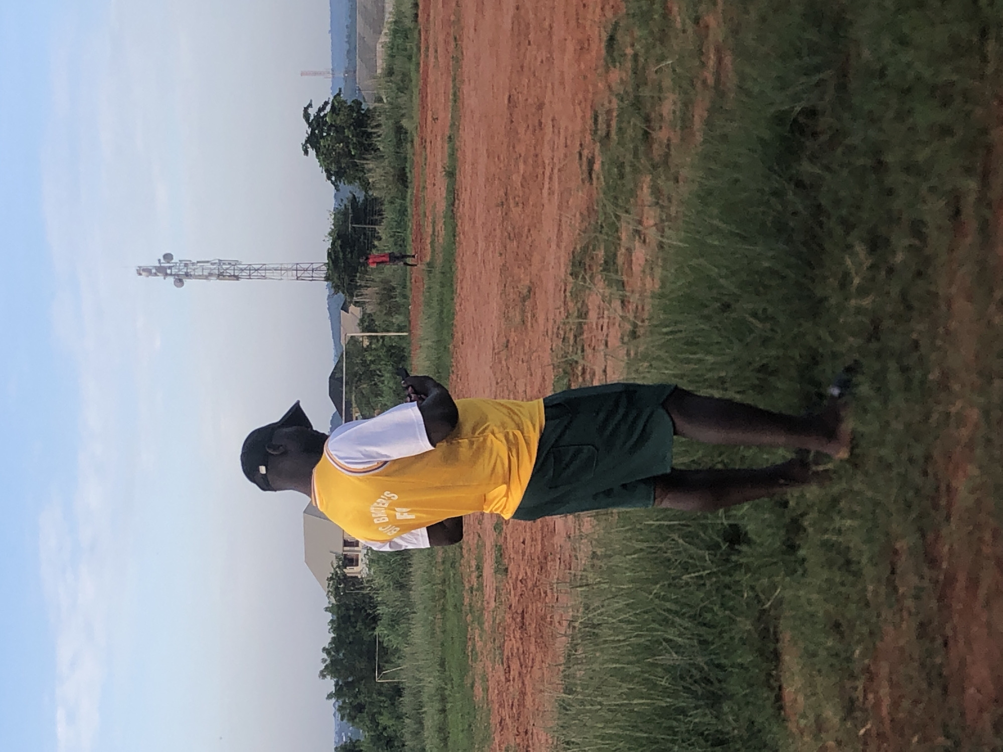 The image depicts an outdoor scene in what appears to be a large, open field, likely a football (soccer) pitch, under a clear, bright daytime sky. The ground consists of sections of reddish-brown earth interspersed with green grass.
In the foreground, a person stands with their back mostly to the camera, looking towards their left. They are wearing a black baseball cap, a yellow sleeveless jersey over a white t-shirt, and dark green shorts. The back of the yellow jersey clearly displays the text "BIG BROTHER'S FC". The person is barefoot and is holding a smartphone in both hands, seemingly engrossed in it.
In the mid-ground, further back on the reddish field, another person is visible wearing a red shirt and dark bottoms. To the left of the main subject, a white goalpost, typical of a soccer field, is partially visible. A tall, metal communication tower with multiple antennas stands prominently in the background. Beyond the field and structures, a landscape of distant hills or mountains can be seen, along with scattered trees and some low-lying buildings.
The scene suggests a casual setting, possibly during or after a sports activity, consistent with the jersey's text. The bright lighting indicates daytime. The reddish soil and type of tower align with the provided location context of Ukwu-Abwa, Nigeria.