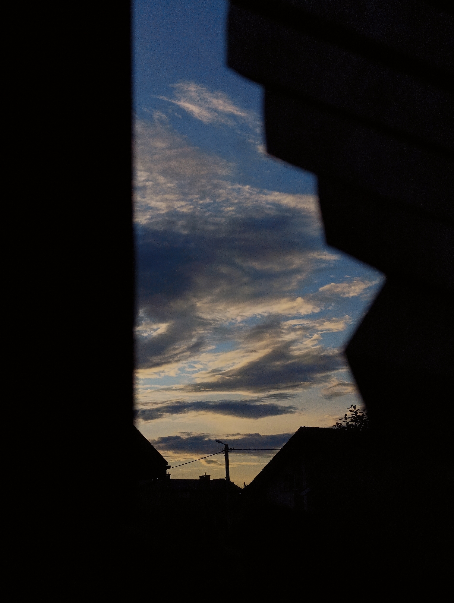 The image shows a silhouetted view of rooftops and a utility pole against a cloudy sky. The foreground is dominated by dark, abstract shapes that frame the scene, suggesting the viewer is looking through a window or an opening. The sky features streaks of white and gray clouds against a backdrop of blue and warm orange hues, indicating either sunrise or sunset. The silhouettes of houses and a single streetlamp are visible on the horizon. No people are visible in the image. The setting appears to be a residential area, possibly in Minsk, Belarus, given the architectural style of the roofs and the presence of a utility pole.